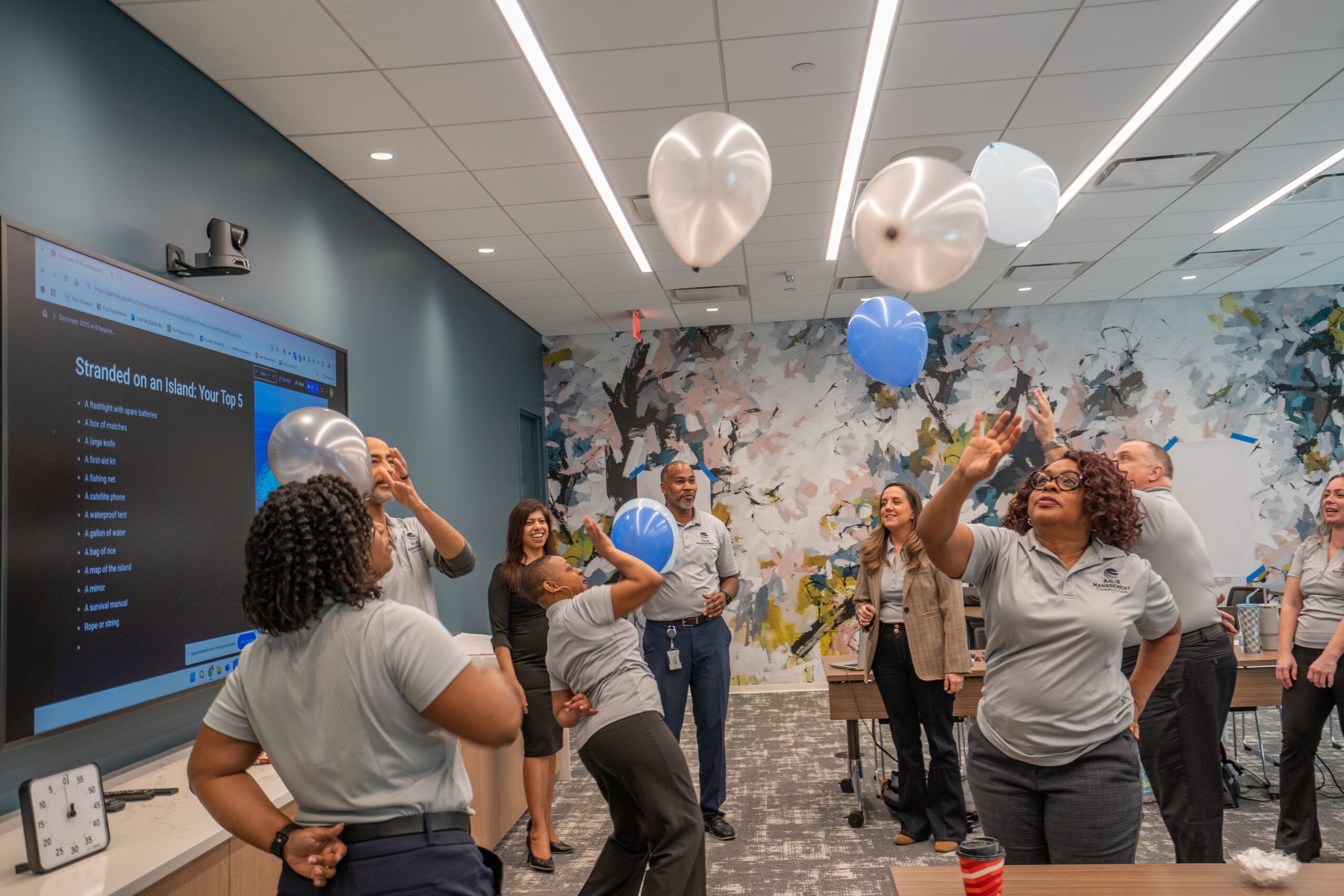Group of people in a room with balloons, some reaching for balloons, and a large screen on the wall.