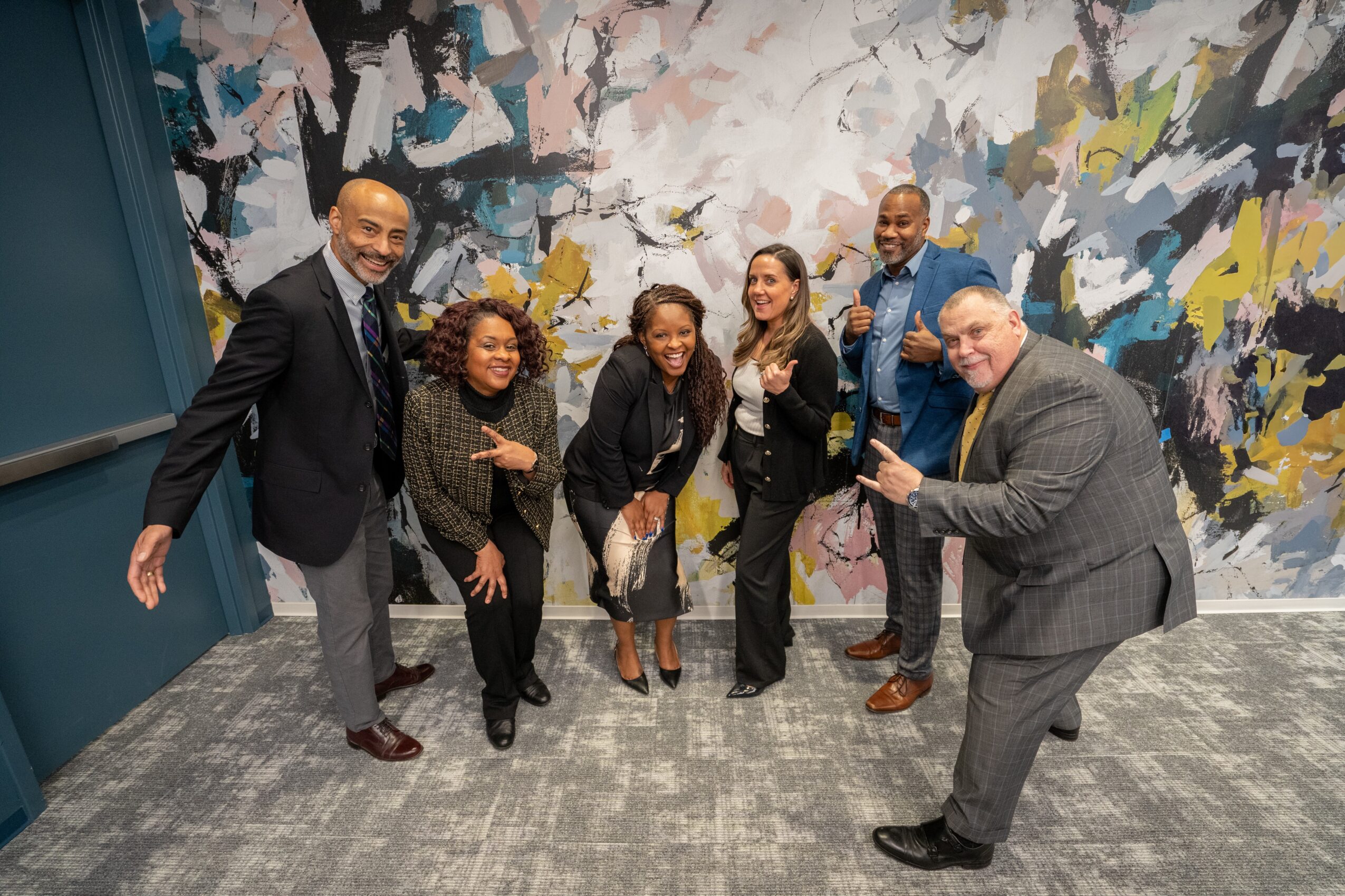 Group of six people standing together indoors, smiling, in front of an abstract wall painting.