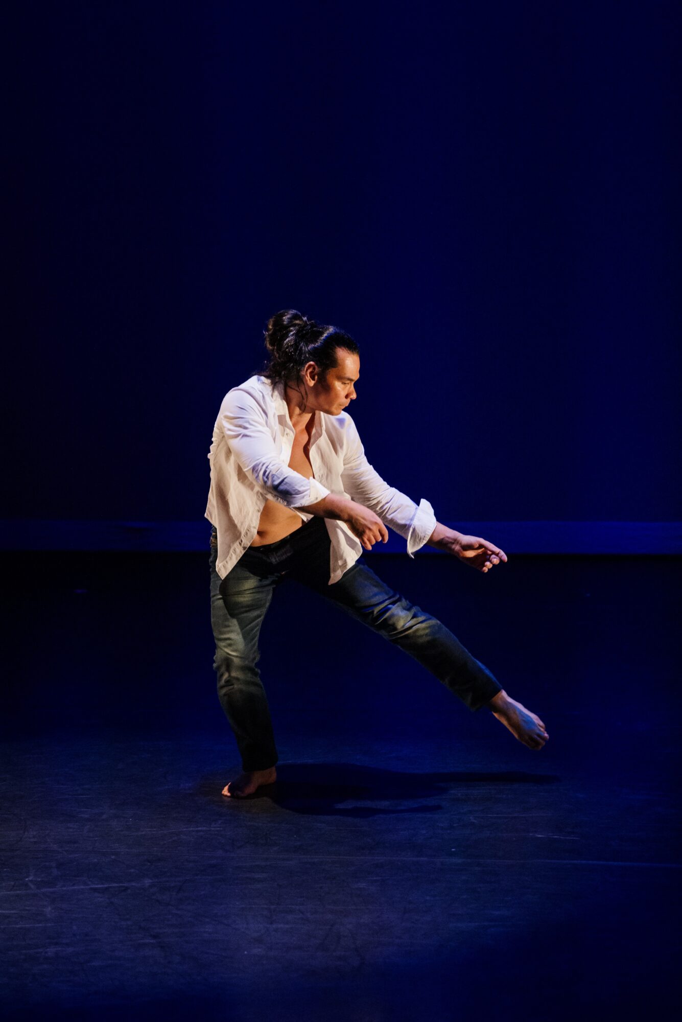 Person dancing on stage with dark background, wearing a white shirt and dark pants, in a dynamic pose.