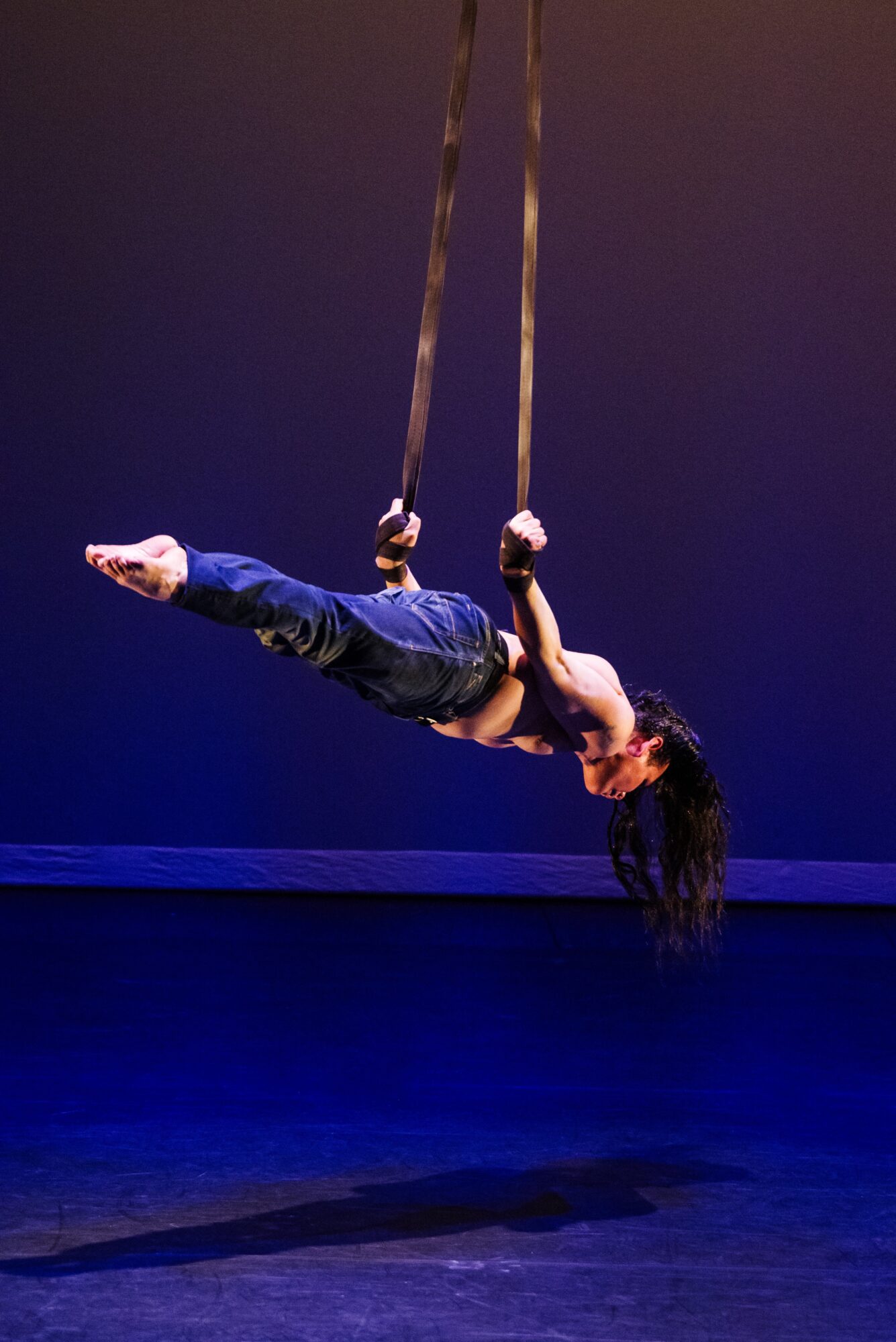 A woman performs aerial acrobatics on ropes, suspended horizontally with her body extended and hair hanging downward.