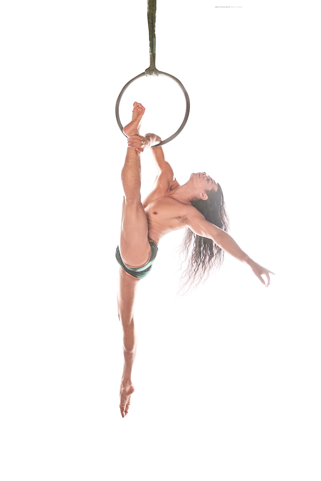 A woman performs an aerial pose on a hoop hanging from the ceiling, with one leg extended upward and arms outstretched.