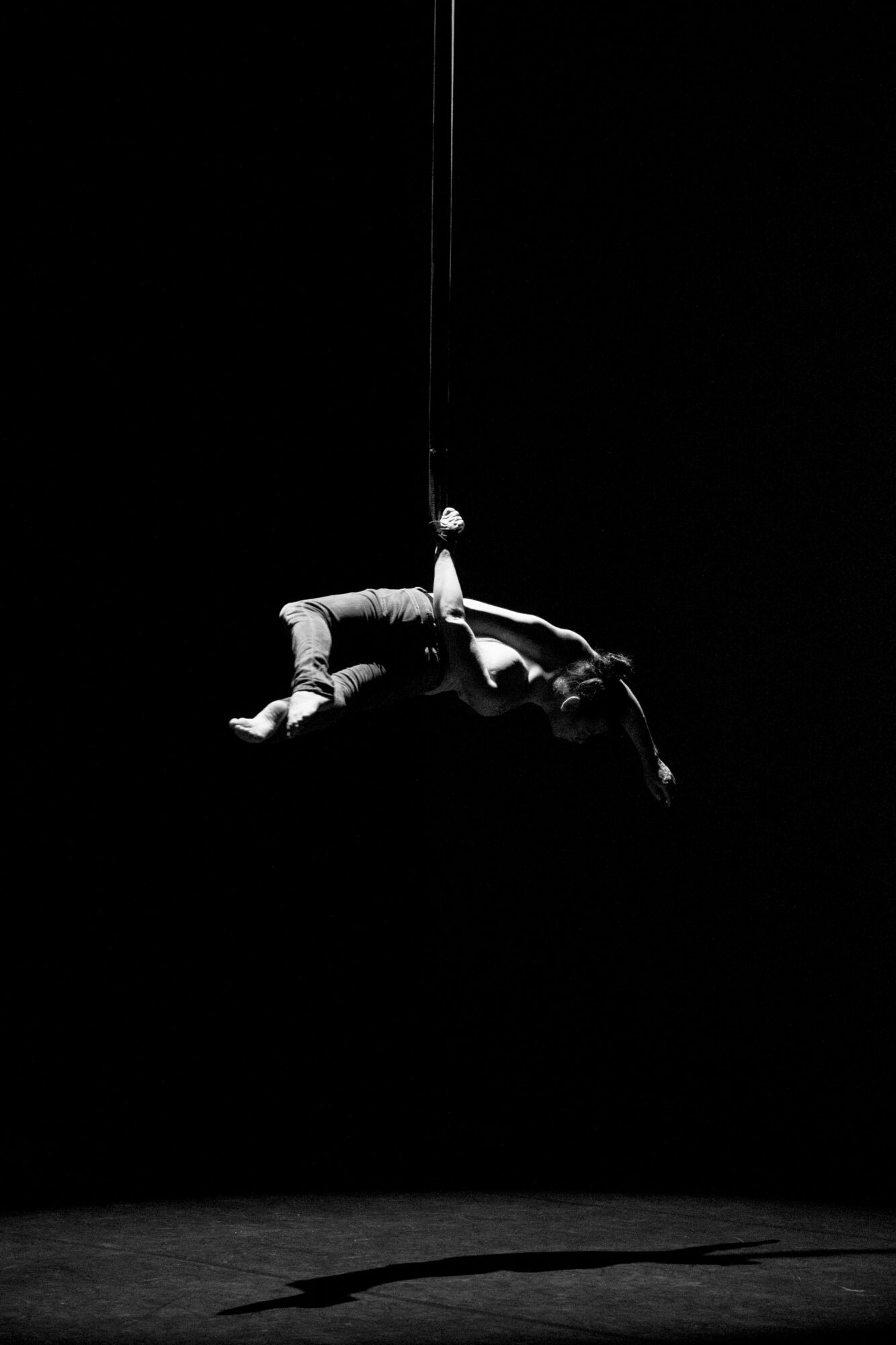 Performer lying on stage with a harness hanging above, shadow cast on the floor, black background, dramatic lighting.