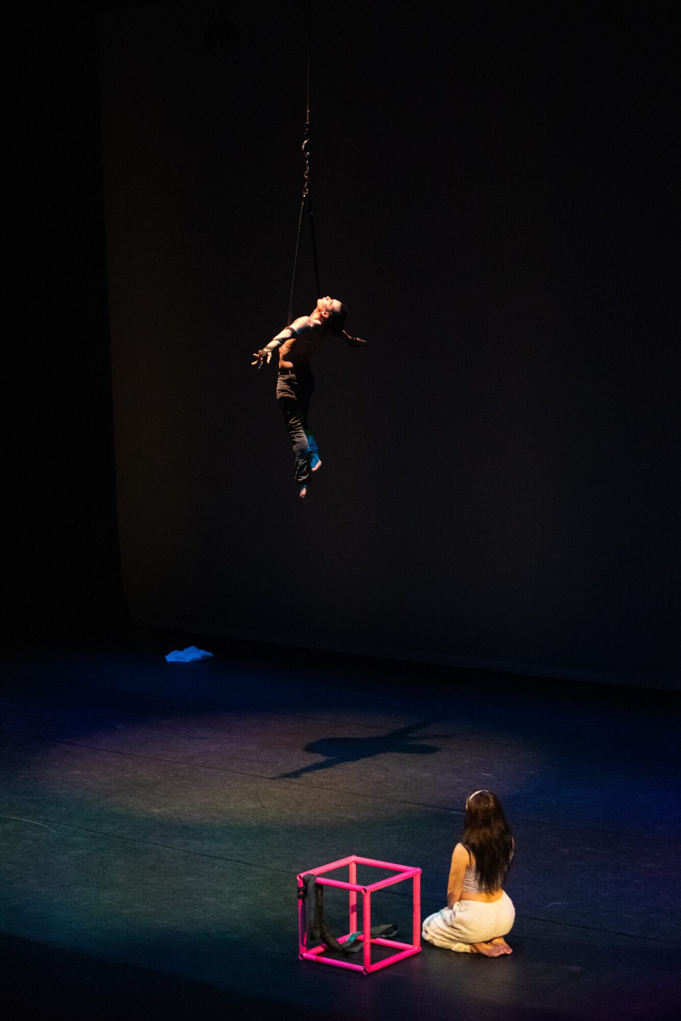 Performer hanging from a rope above a person sitting on the stage, with a pink cube nearby, in a dark theater.