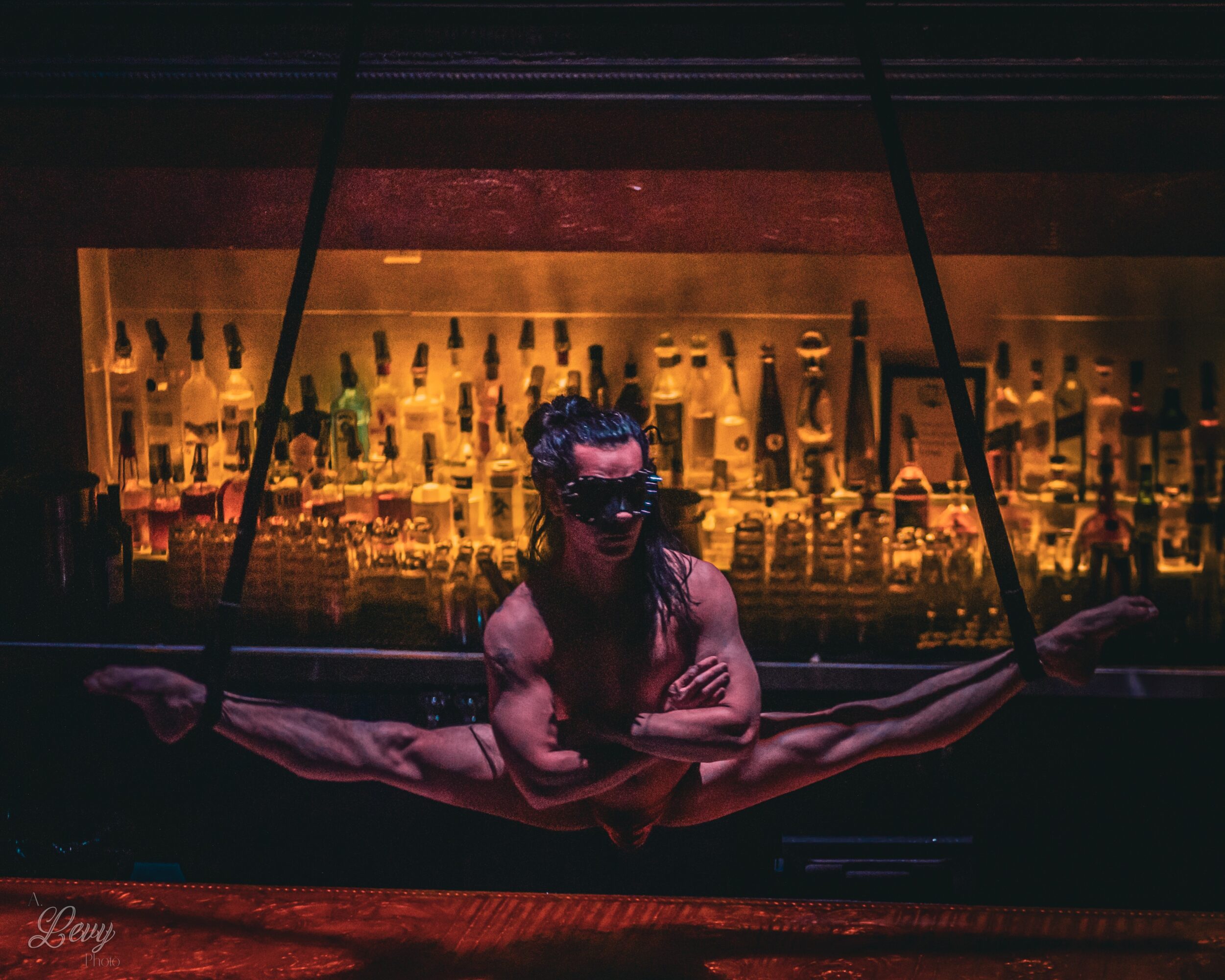 Person with makeup and costume performing a split in front of a bar with bottles, holding ropes on each side.