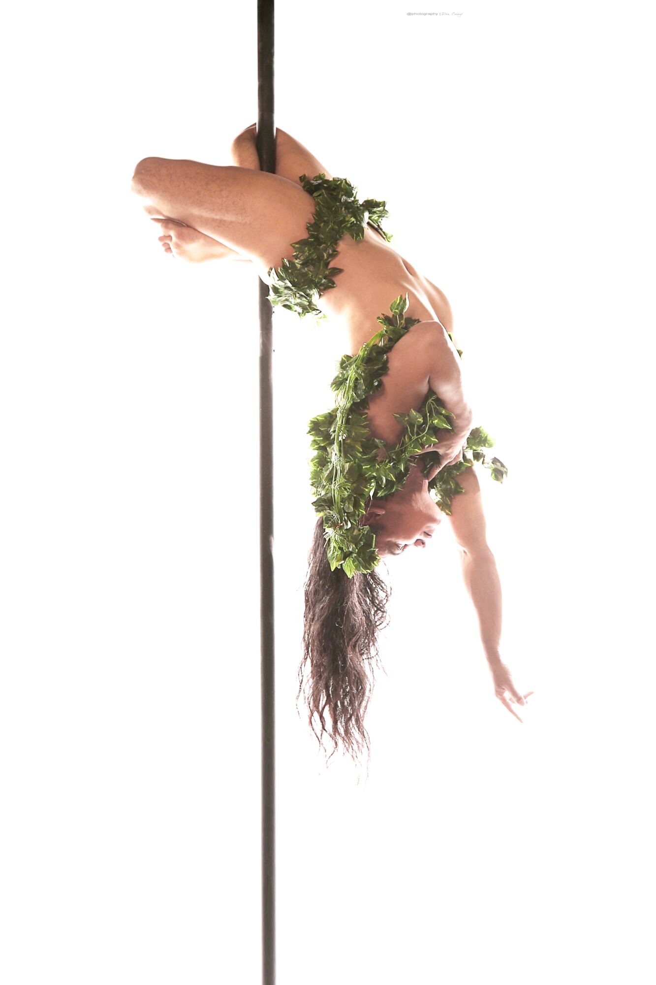 Person hanging upside down from a pole, covered with green leaves, with long hair hanging downward.