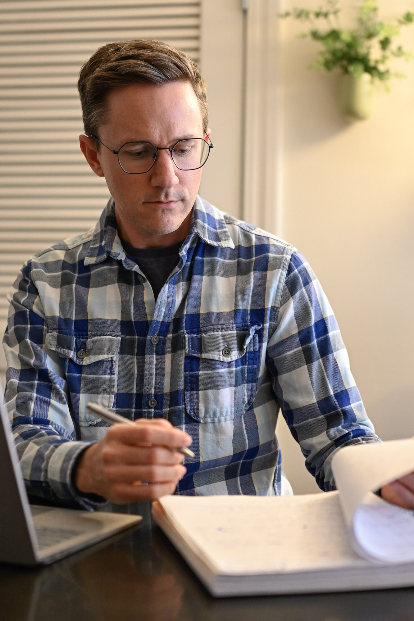 Man with glasses and plaid shirt looking down at open book on table.