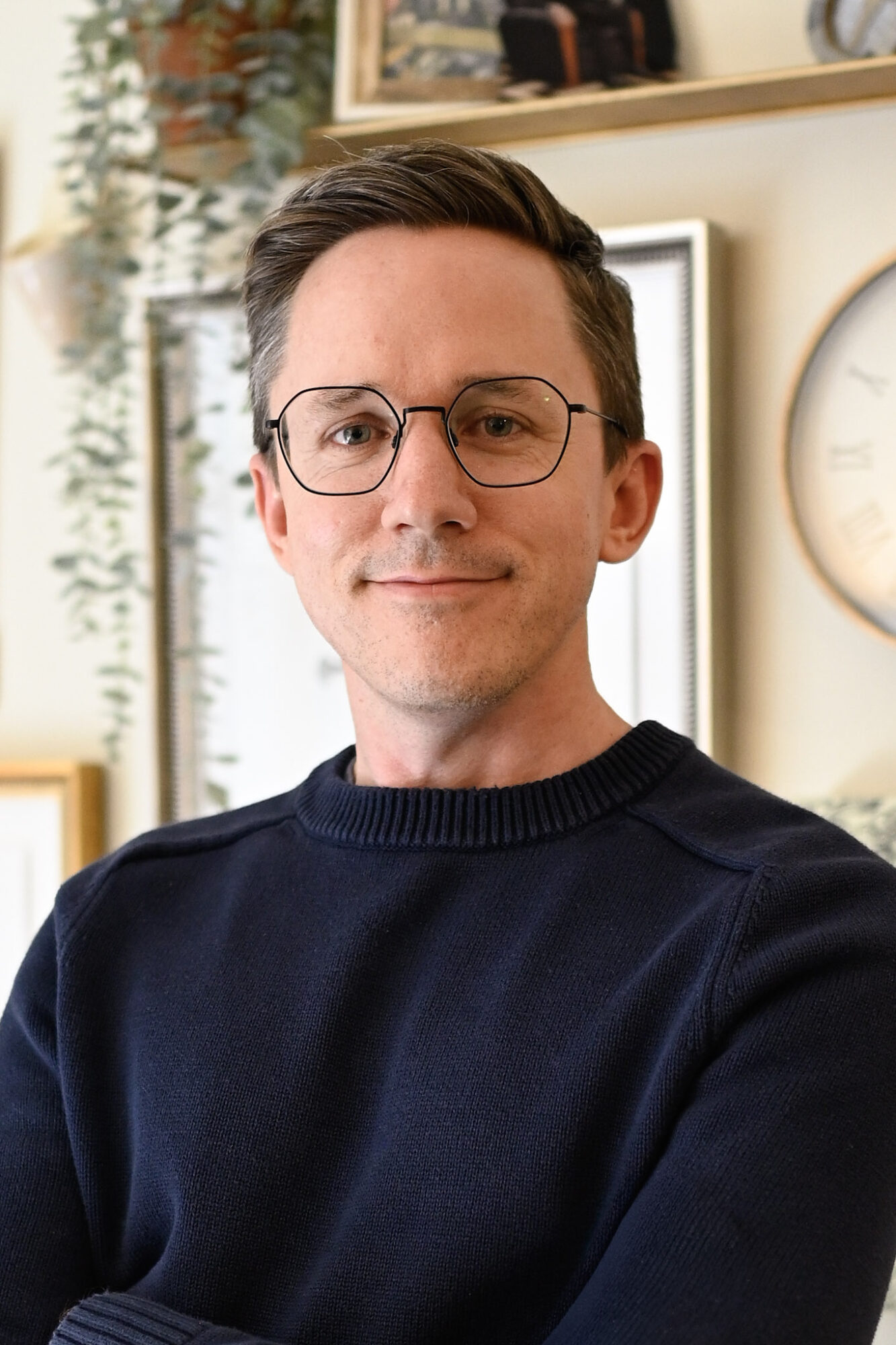 Man with glasses and short dark hair smiling indoors, wearing a dark sweater, in a room with shelves and a clock.