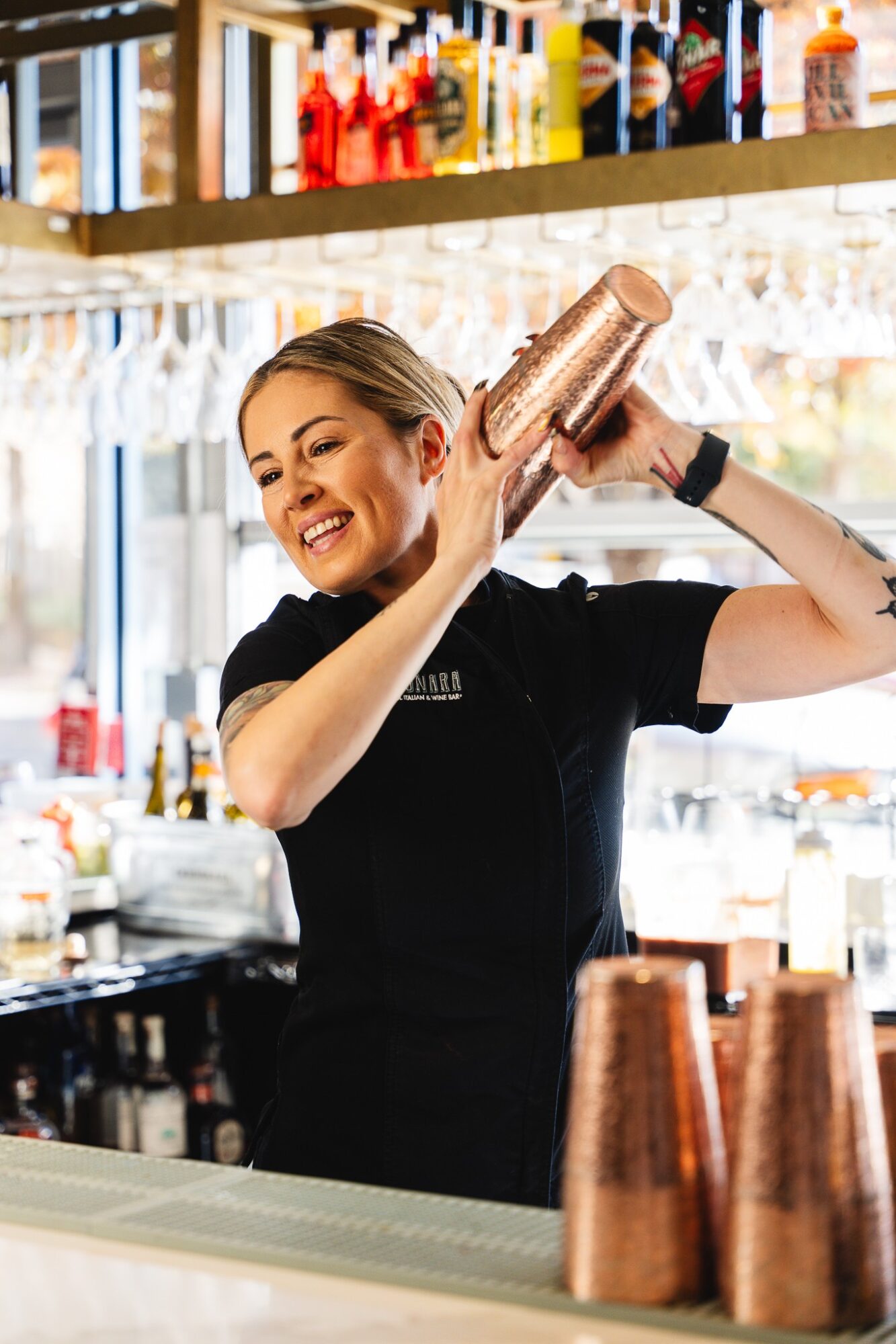 Bartender smiling and shaking a cocktail shaker behind bar counter with bottles on shelf above.