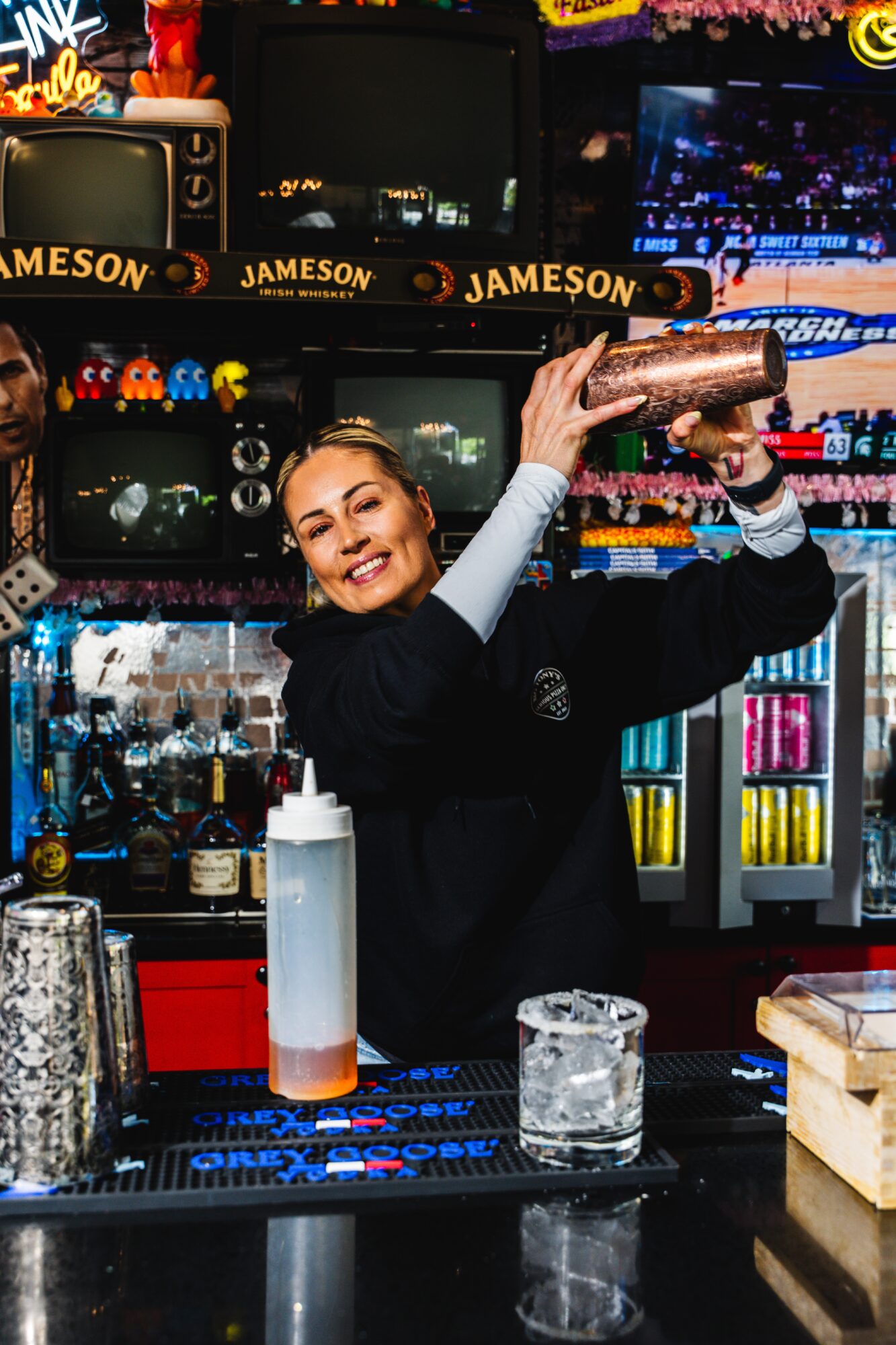 Bartender smiling and shaking a cocktail shaker behind bar with colorful bottles and glasses.