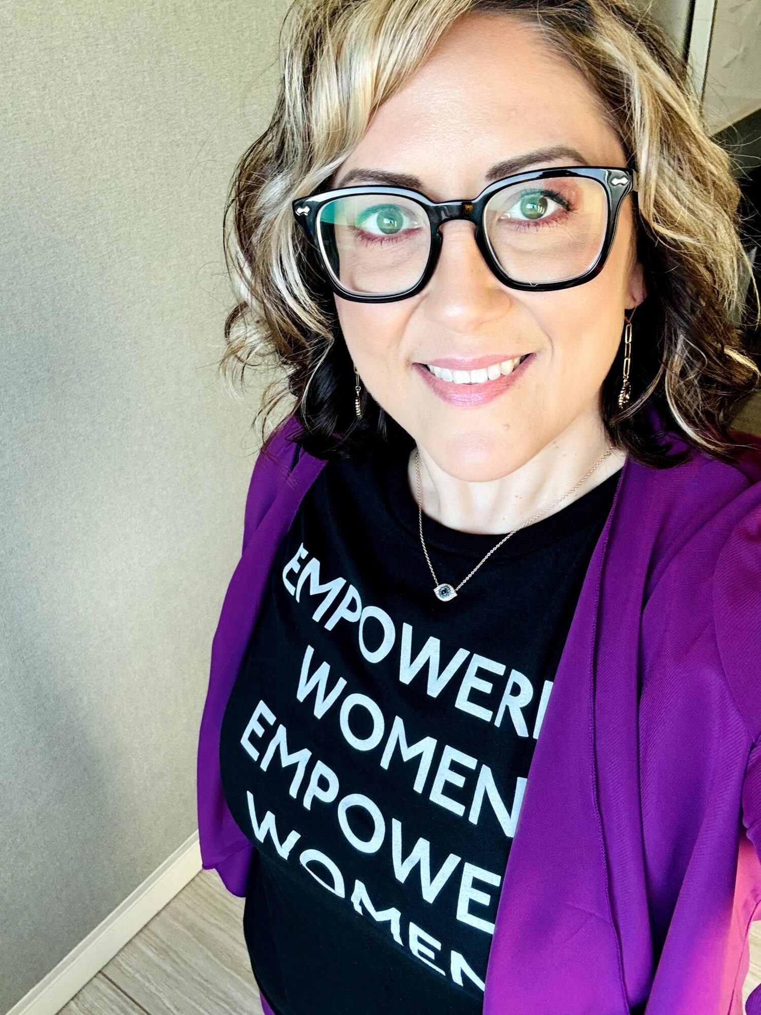 Woman with glasses and wavy hair smiling, wearing a black shirt with white text and a purple jacket.