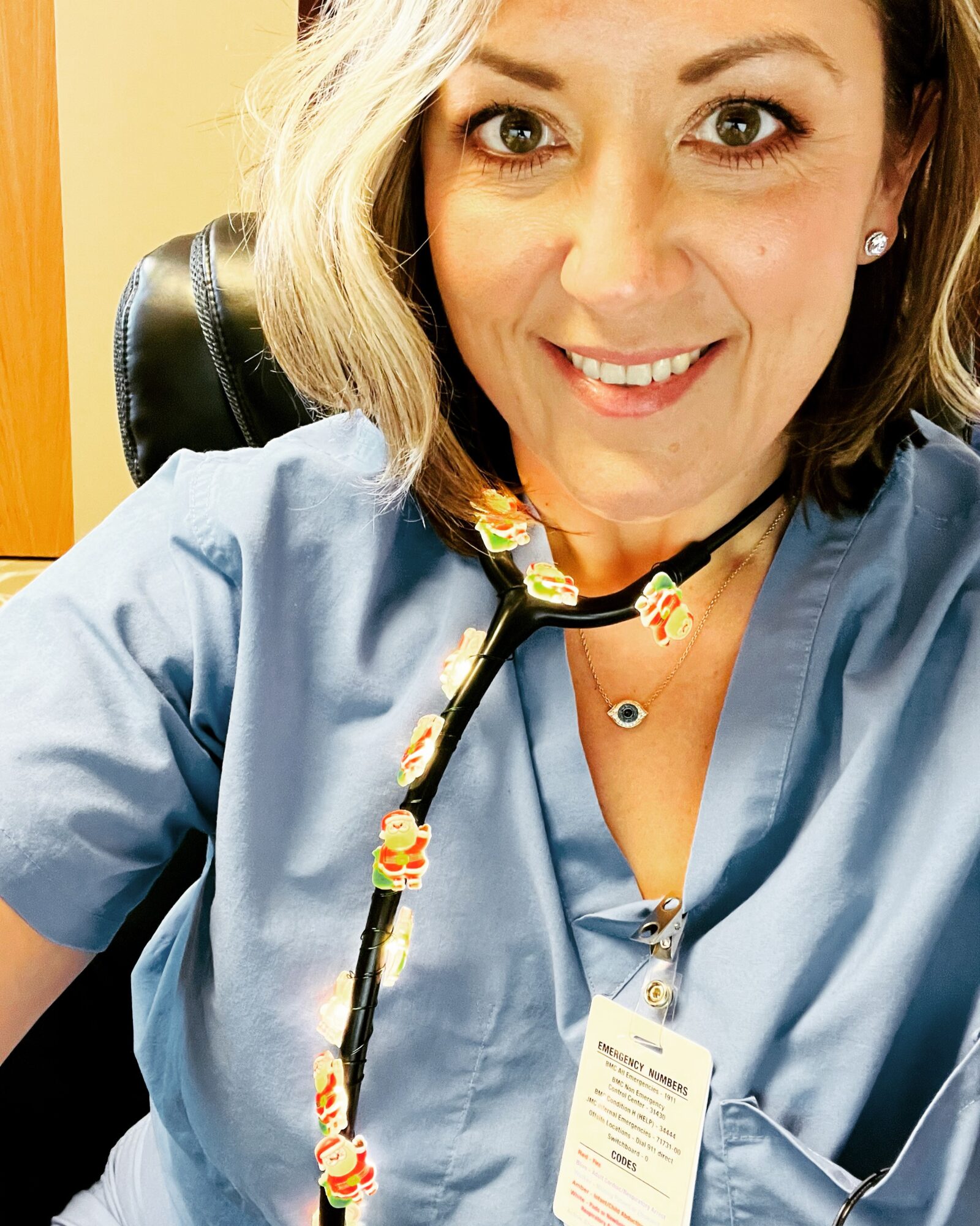 Woman with blonde hair smiling, wearing blue scrubs and a colorful lanyard, sitting in an office chair.