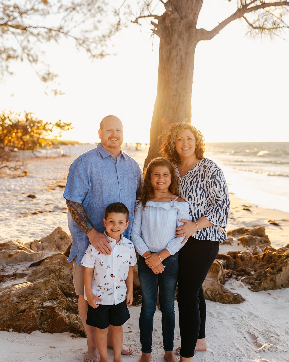 Family of four standing on beach near tree, smiling, with ocean in background during sunset.