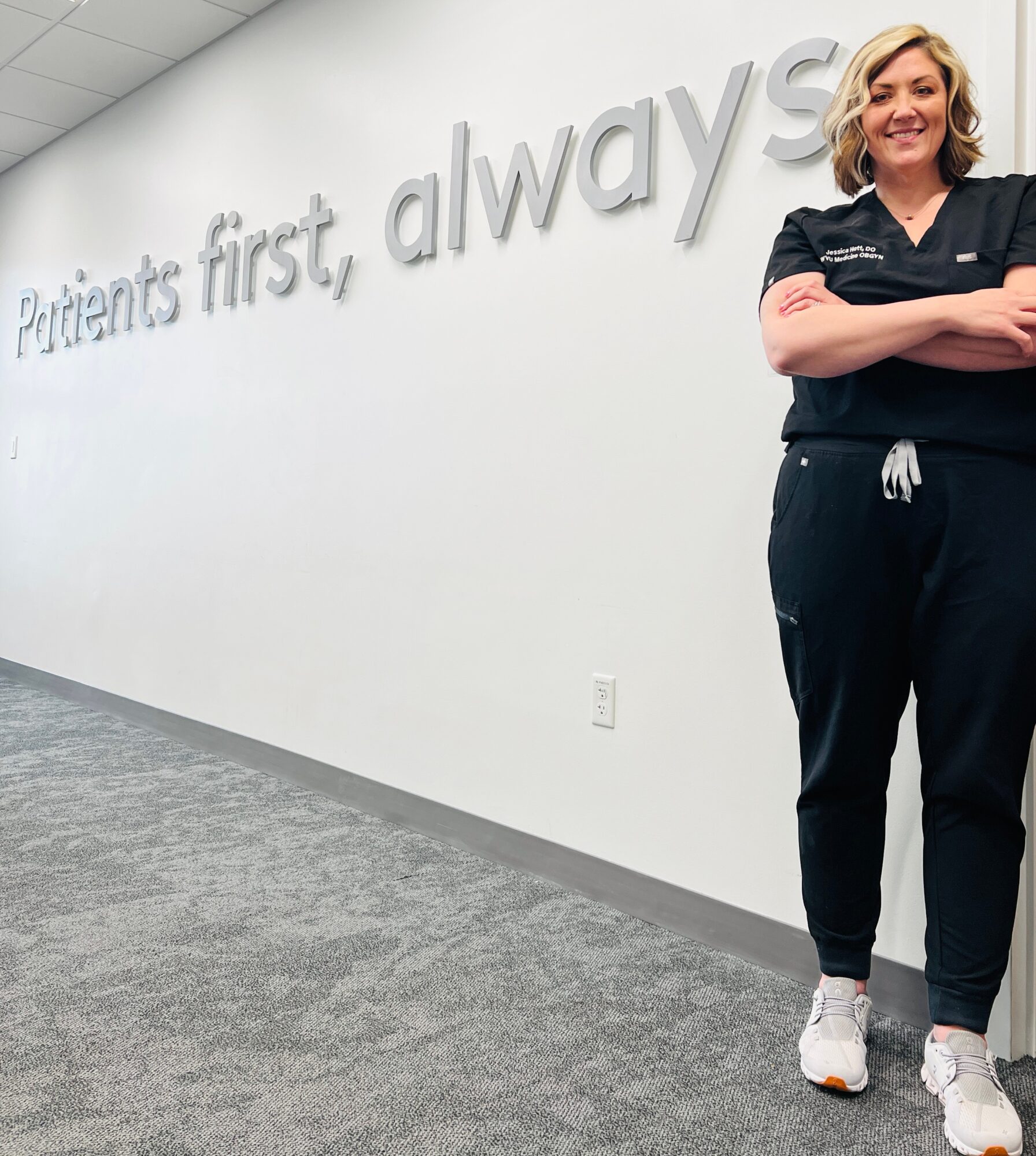 A woman in medical scrubs standing with arms crossed in front of a wall with the words 'Patients first, always'.
