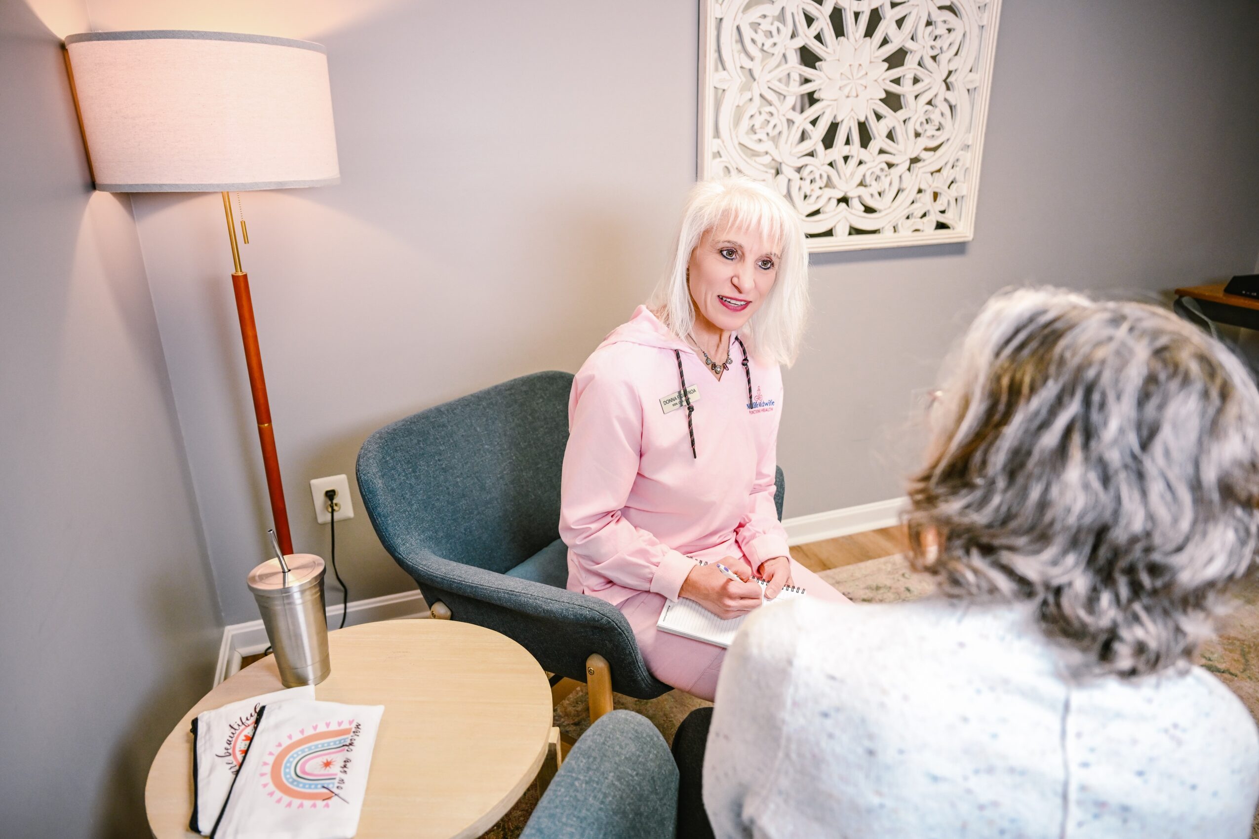 Two women sitting in a room, one facing the camera with white hair and pink shirt, the other with gray hair, back to camera.