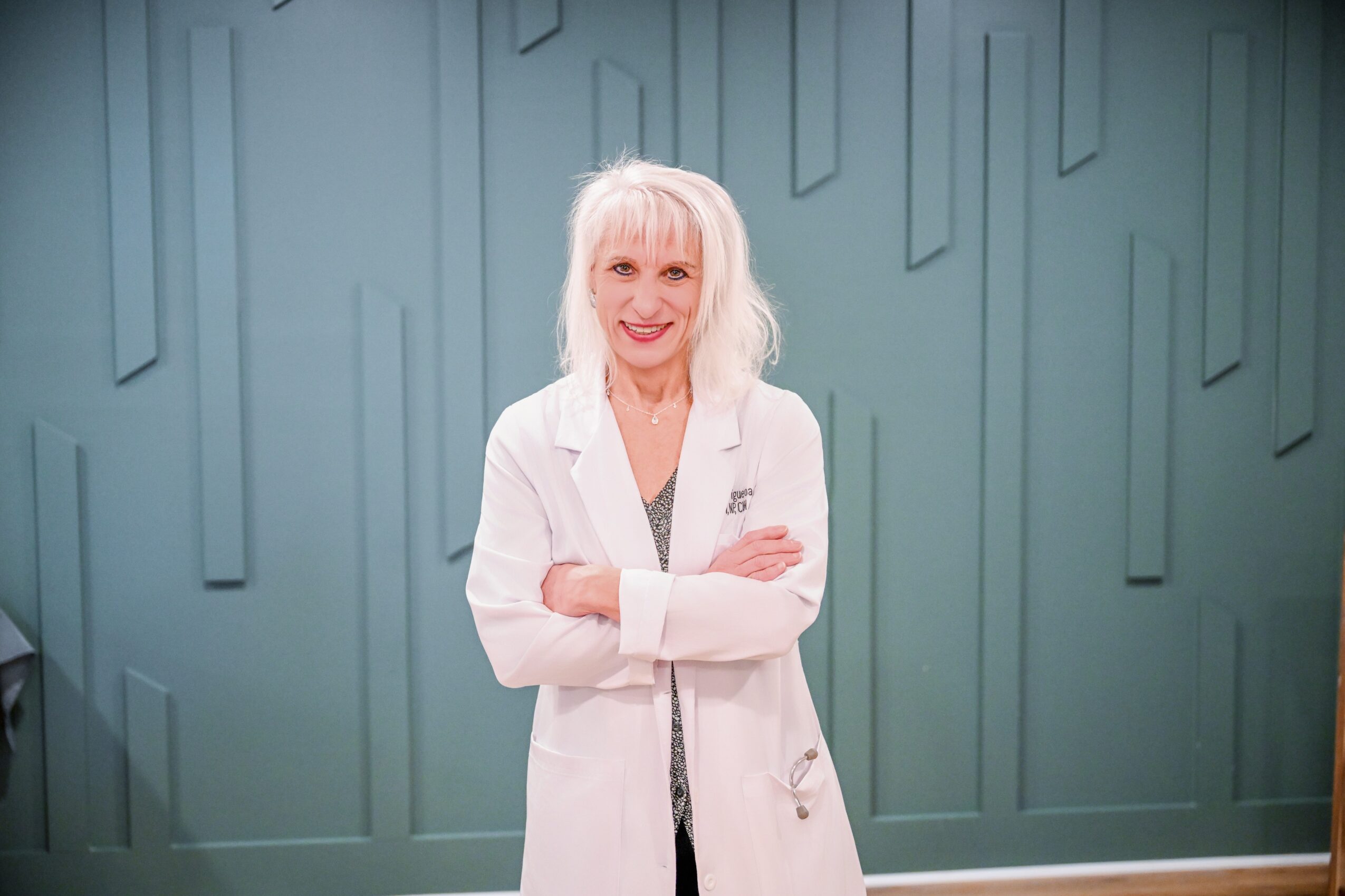 Older woman with white hair smiling, wearing a white coat, standing in front of a patterned teal wall.