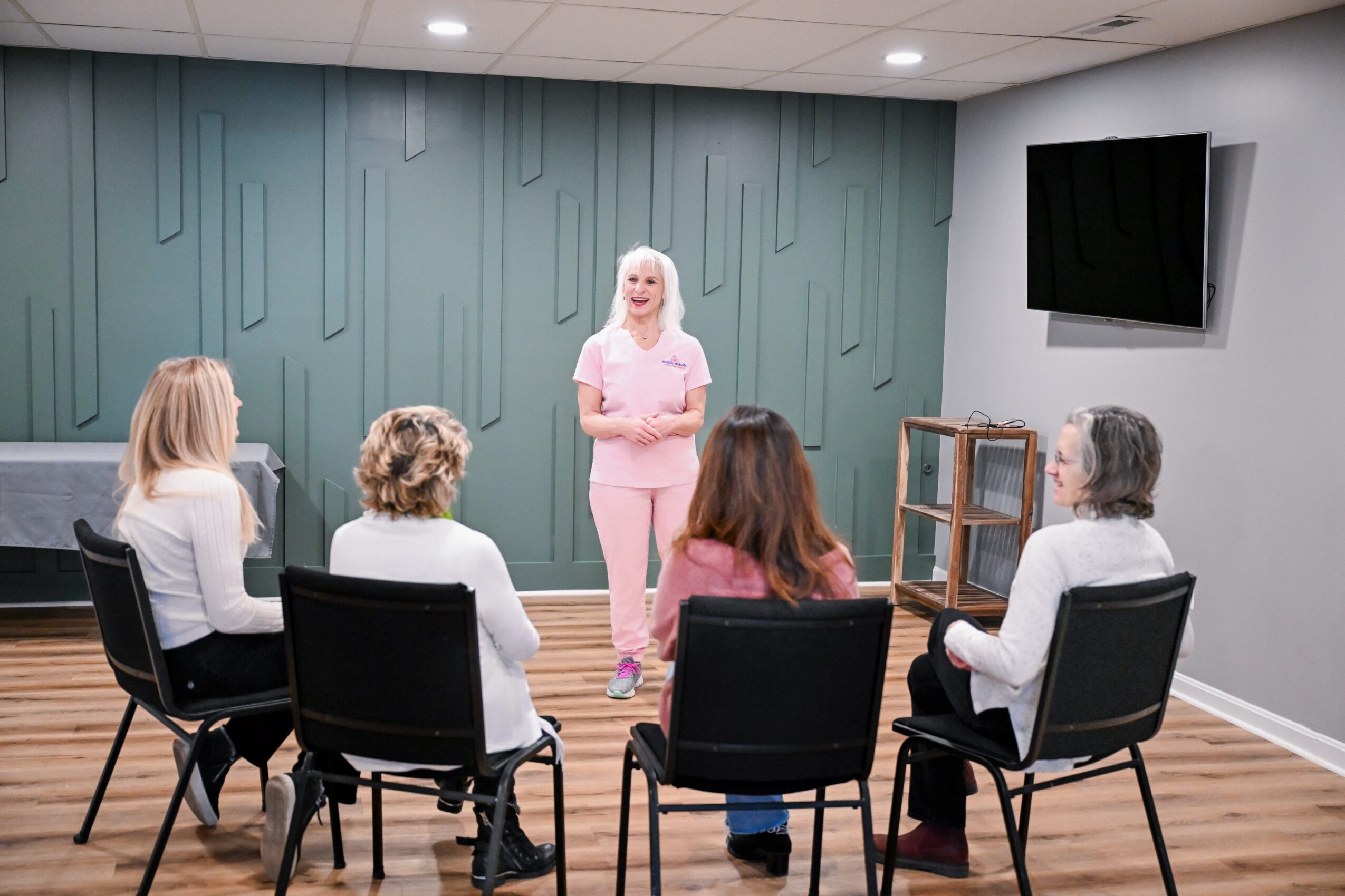 A woman in pink scrubs stands in front of four seated women in a room with a green wall and wooden floor, watching her.