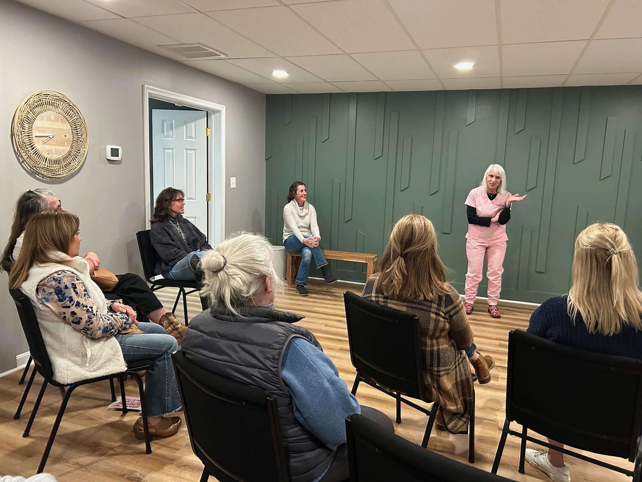 Group of people seated in a room listening to a woman speaking, with a green wall and wooden floor.