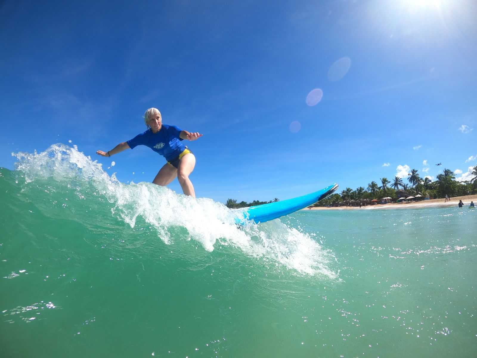 Person surfing on a wave with a clear blue sky and trees in the background, sunlight shining brightly.