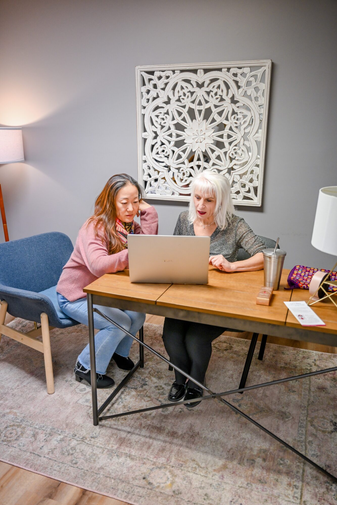Two women sit at a wooden table looking at a laptop, with a decorative wall art behind them.