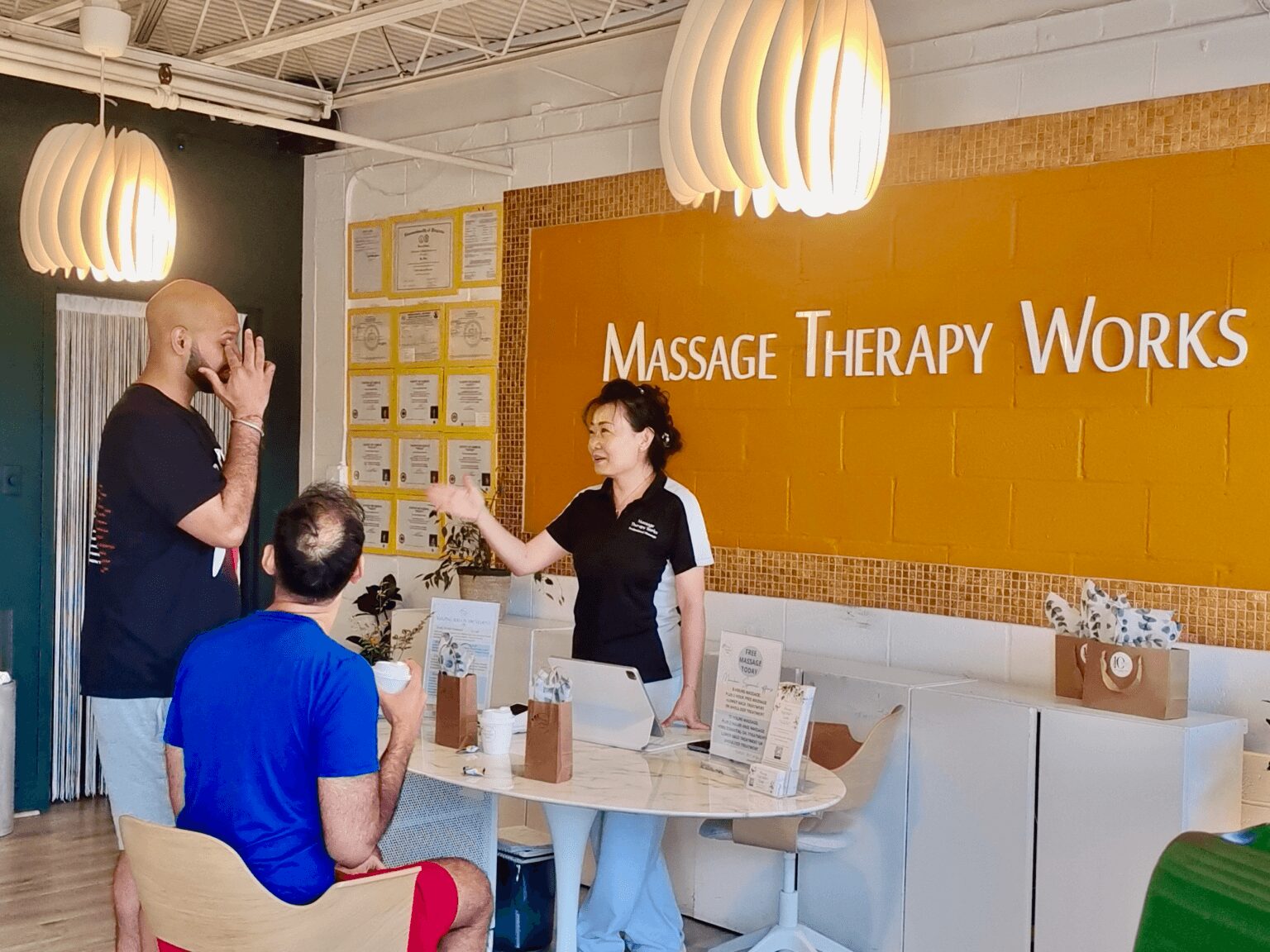 A massage therapy reception area with three people, a woman behind the counter, and two men talking, in a modern space.