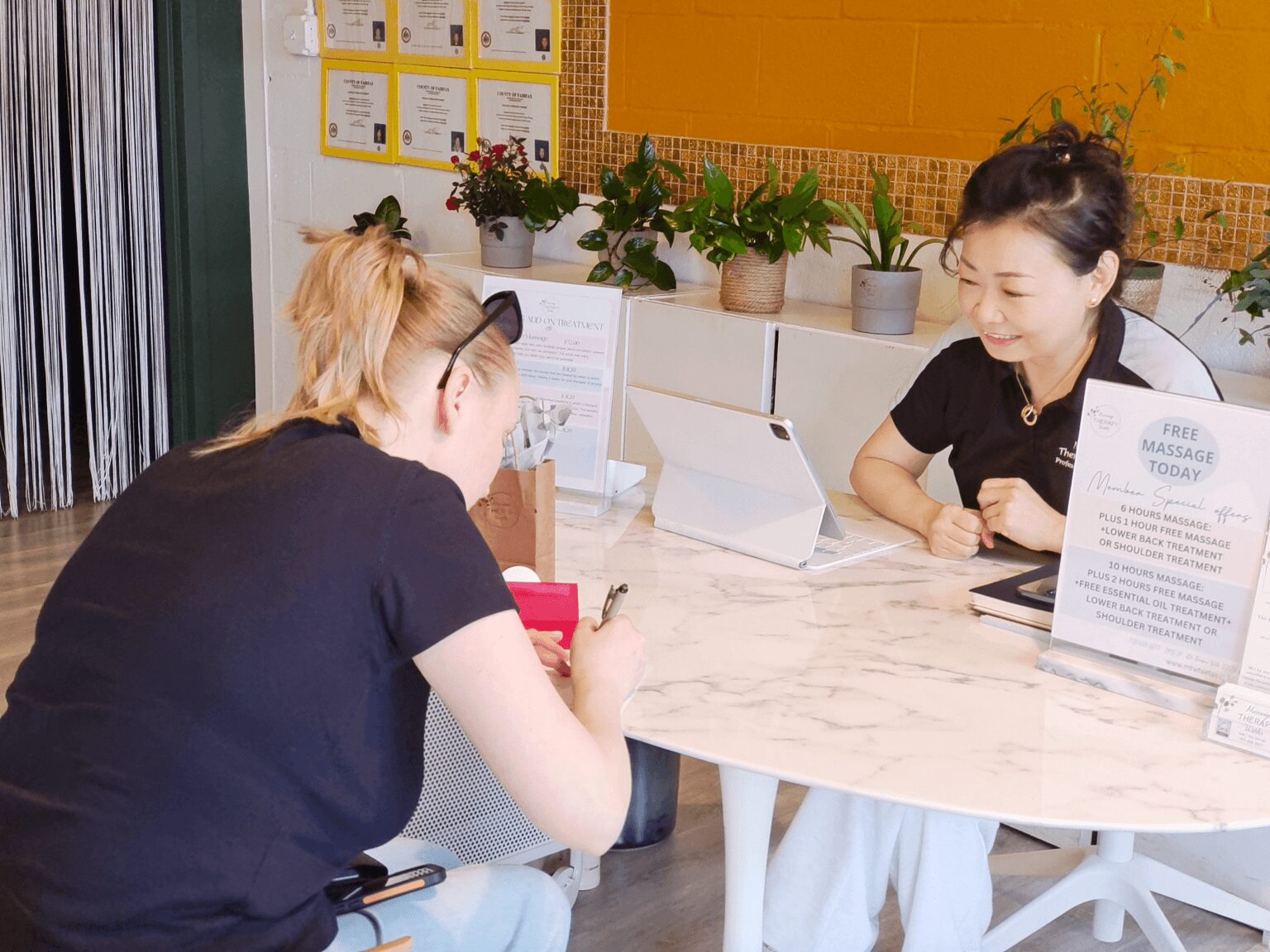 Two women sit at a round marble table, engaging in conversation, with plants and informational signs in the background.