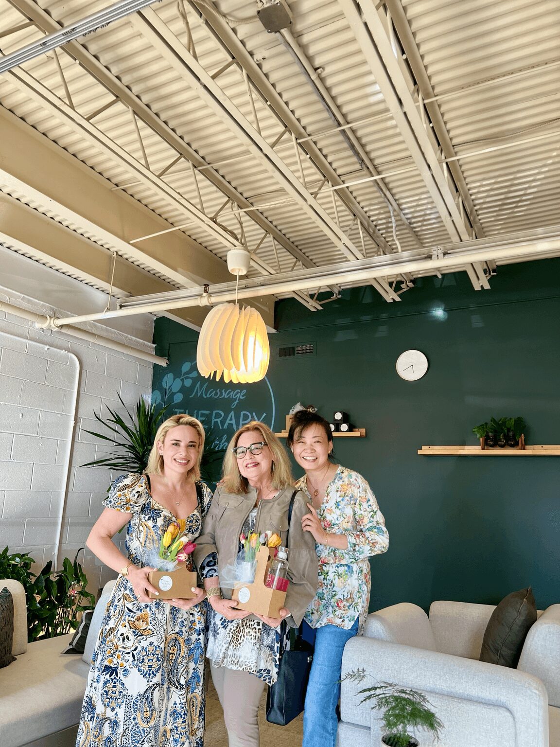 Three women standing indoors, holding drinks and smiling, with a dark green wall, a shelf, and a sofa in the background.