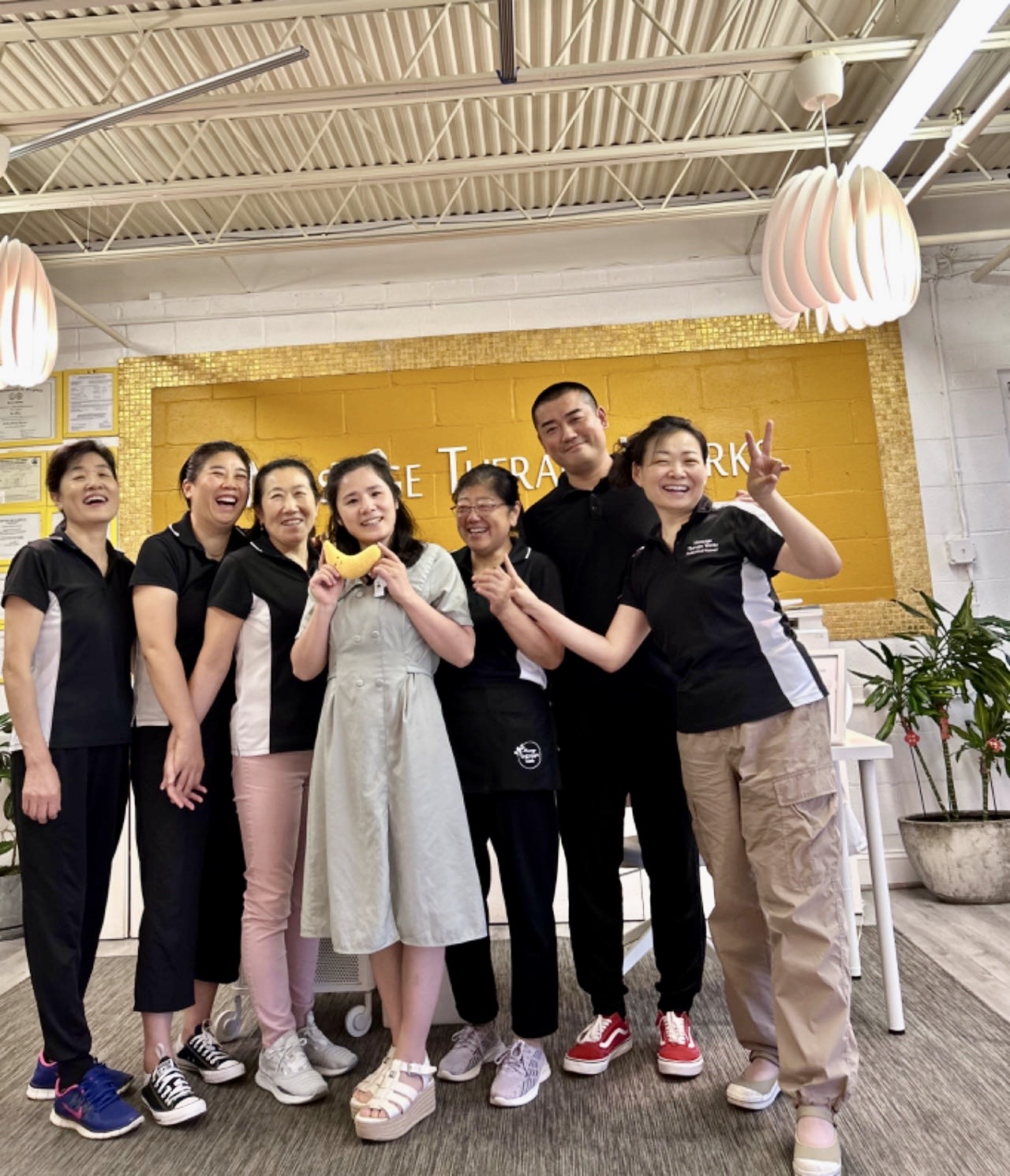 Group of seven people smiling and posing indoors, some making peace signs, in front of a yellow wall with text, with hanging lights and plants nearby.