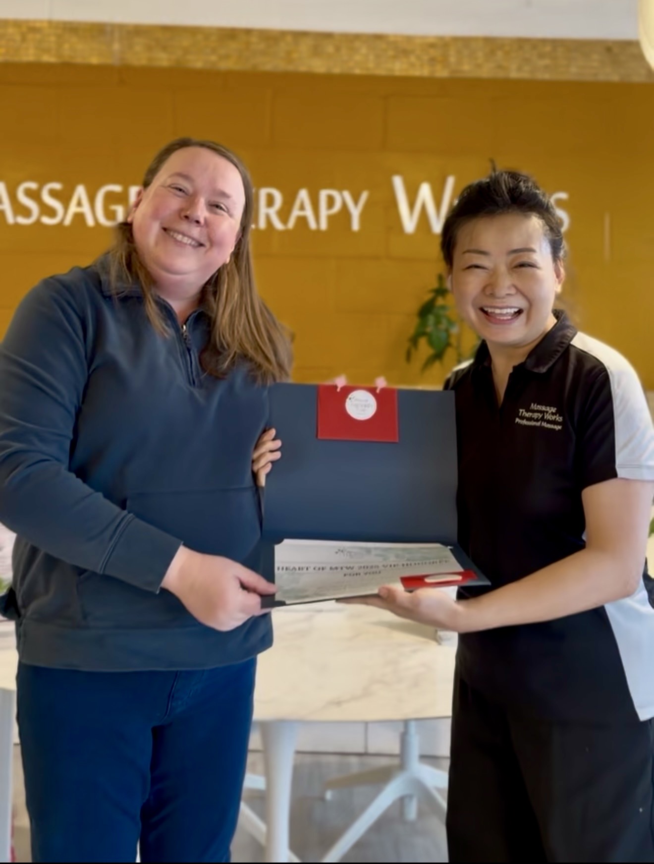 Two women smiling and holding a certificate and a folder in a wellness center, with a yellow wall and green plant behind them.