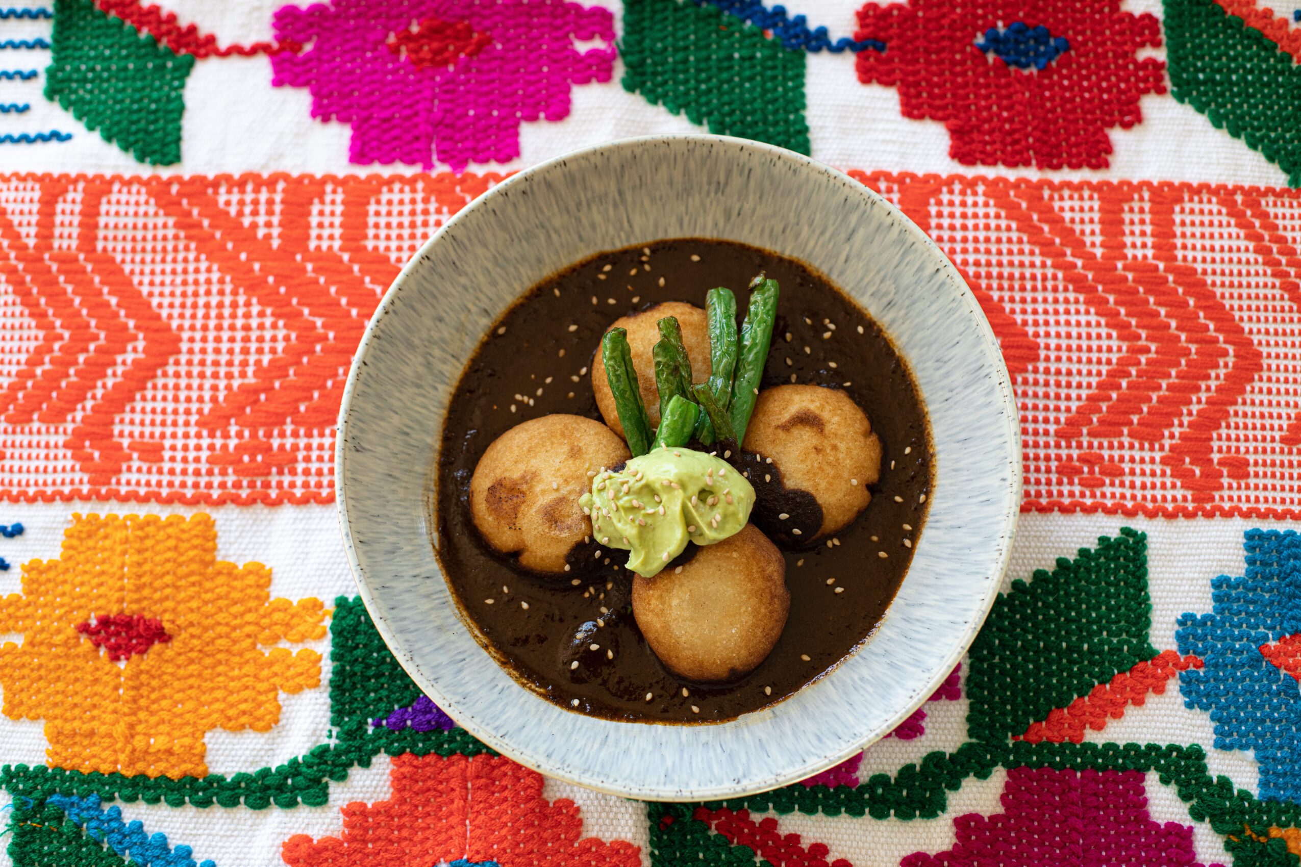 Bowl of dark soup with four round dumplings, green beans, and a dollop of green sauce, on colorful embroidered cloth