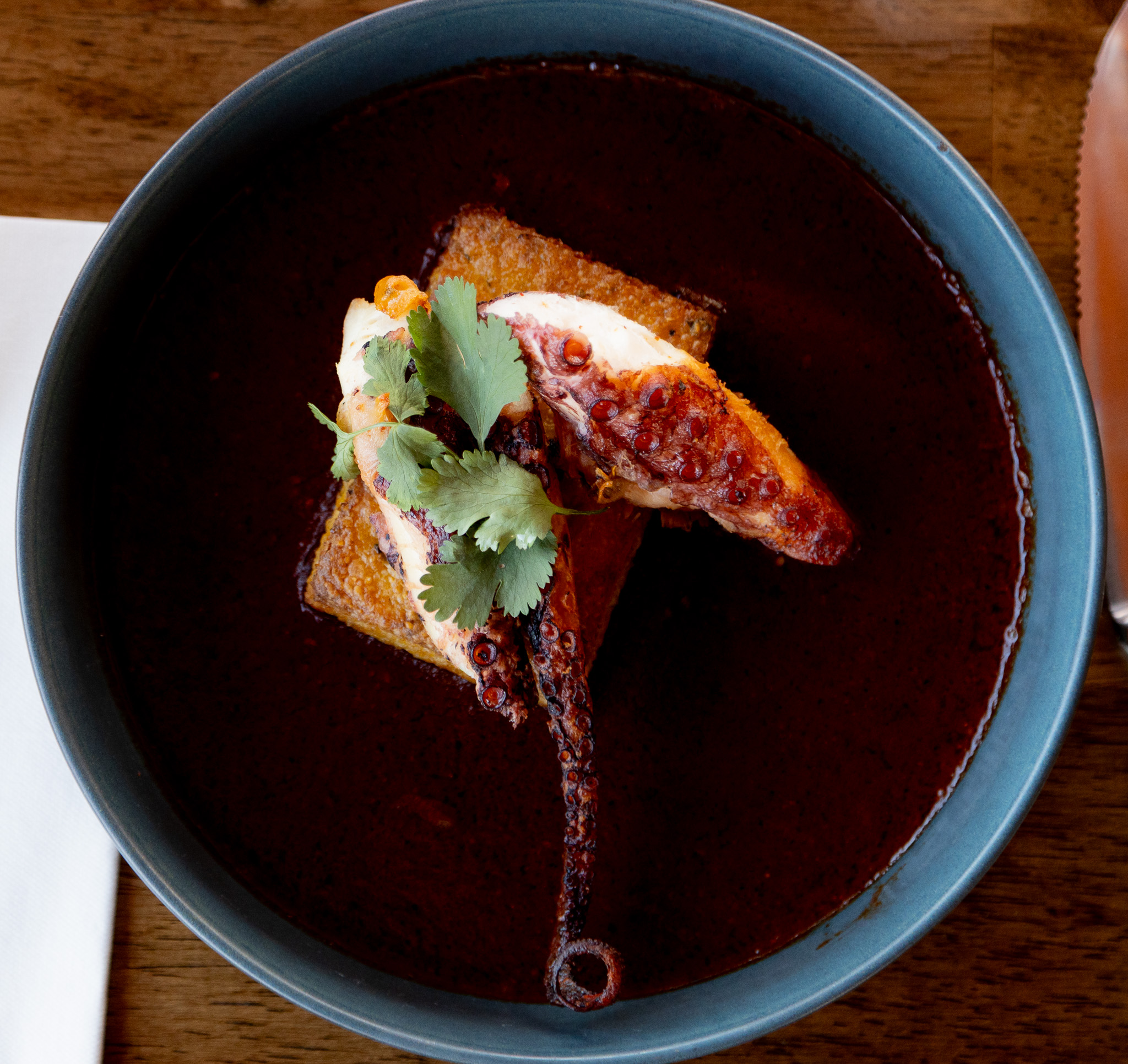 Bowl of dark soup with a poached egg, toast, and cilantro garnish, on a wooden table.
