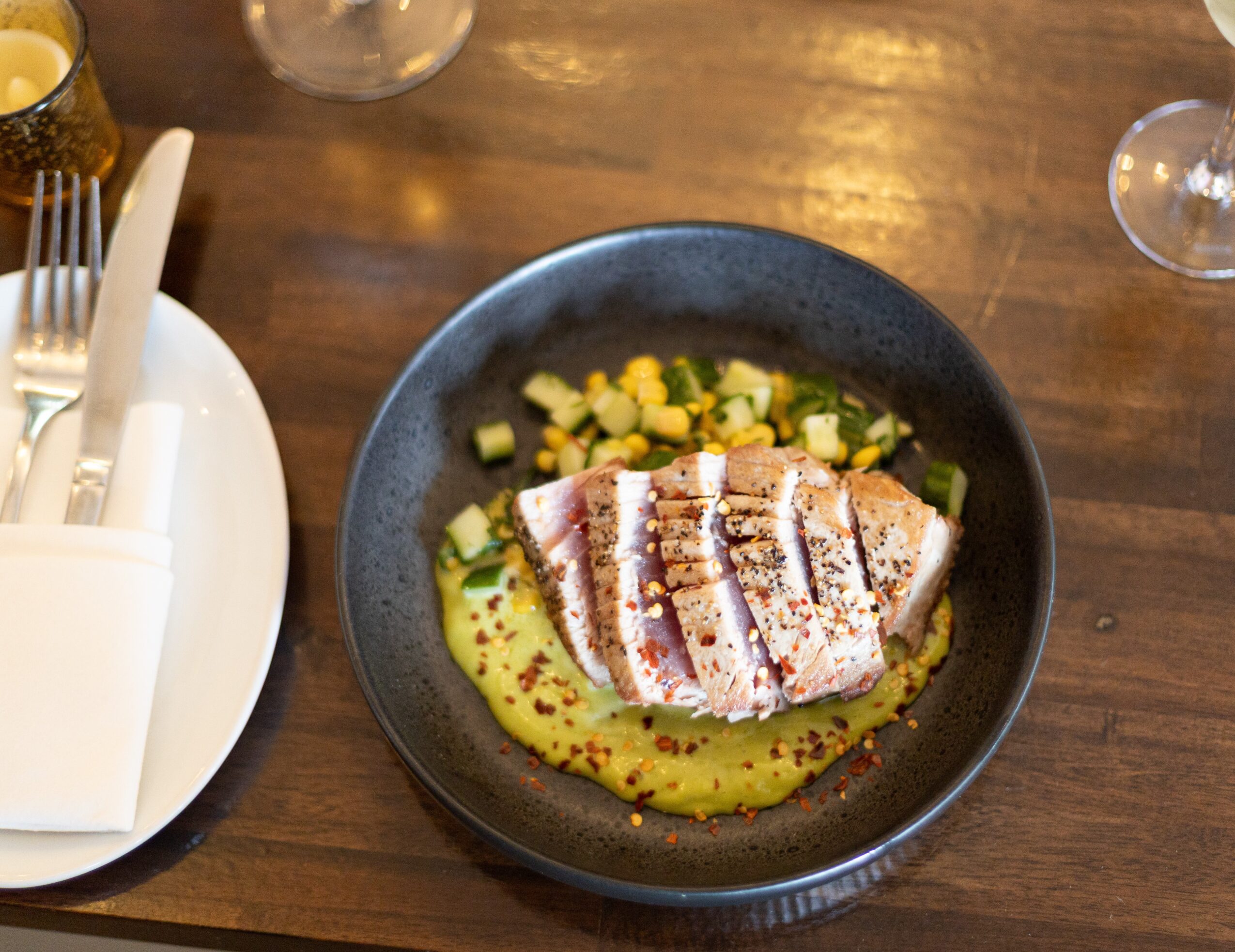 Sliced seared tuna with green sauce and chopped vegetables on a black plate, on a wooden table.