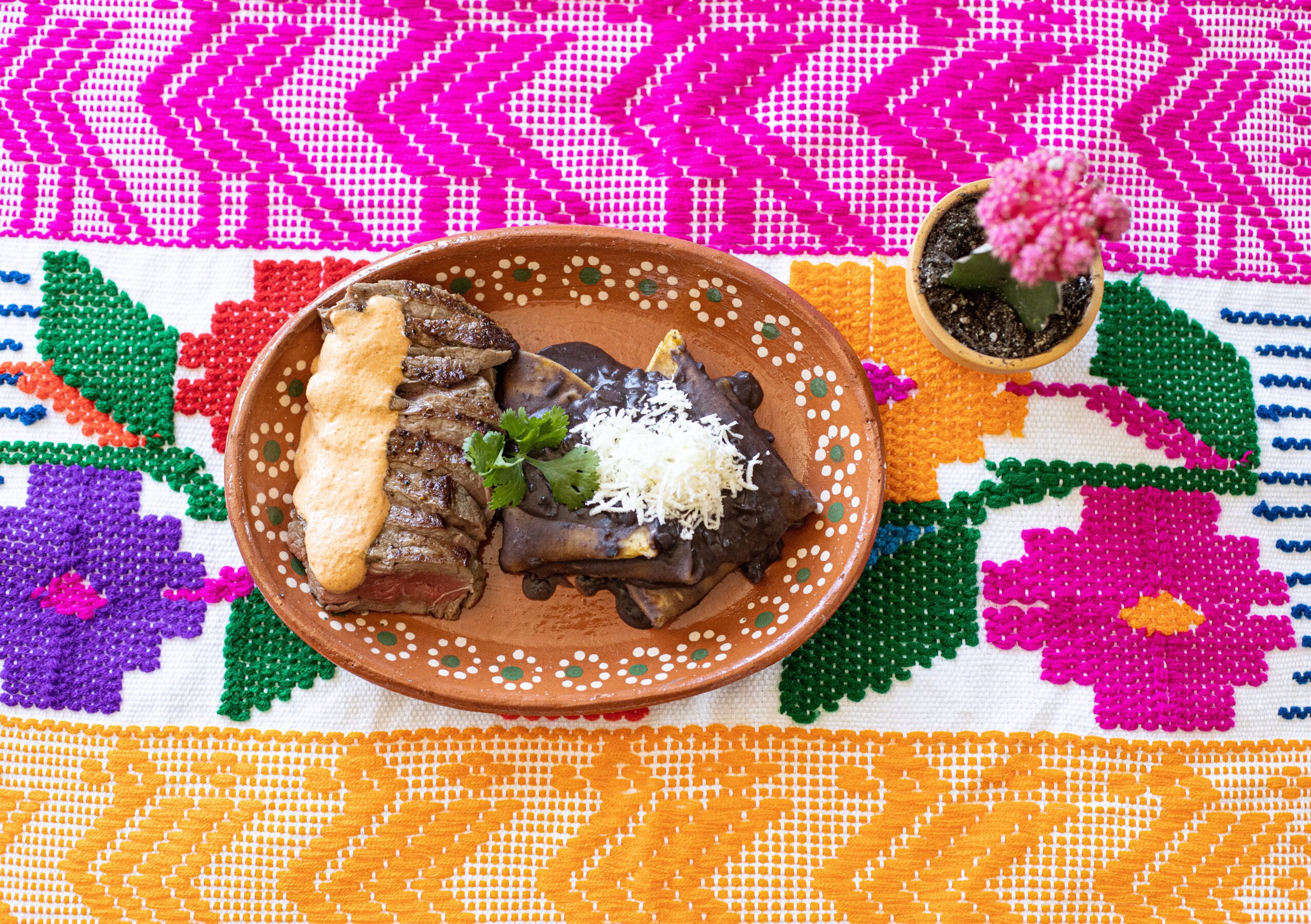 Plate with meat, rice, and a fried item on a colorful embroidered tablecloth, with a small potted pink flower nearby.
