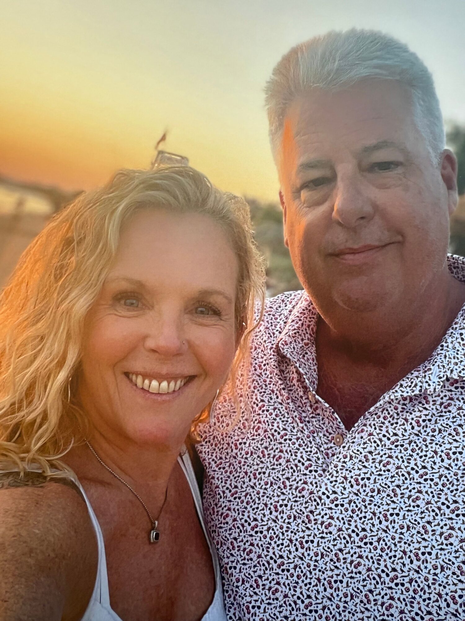 Two people smiling outdoors during sunset, one older man and one woman with curly hair, close together.