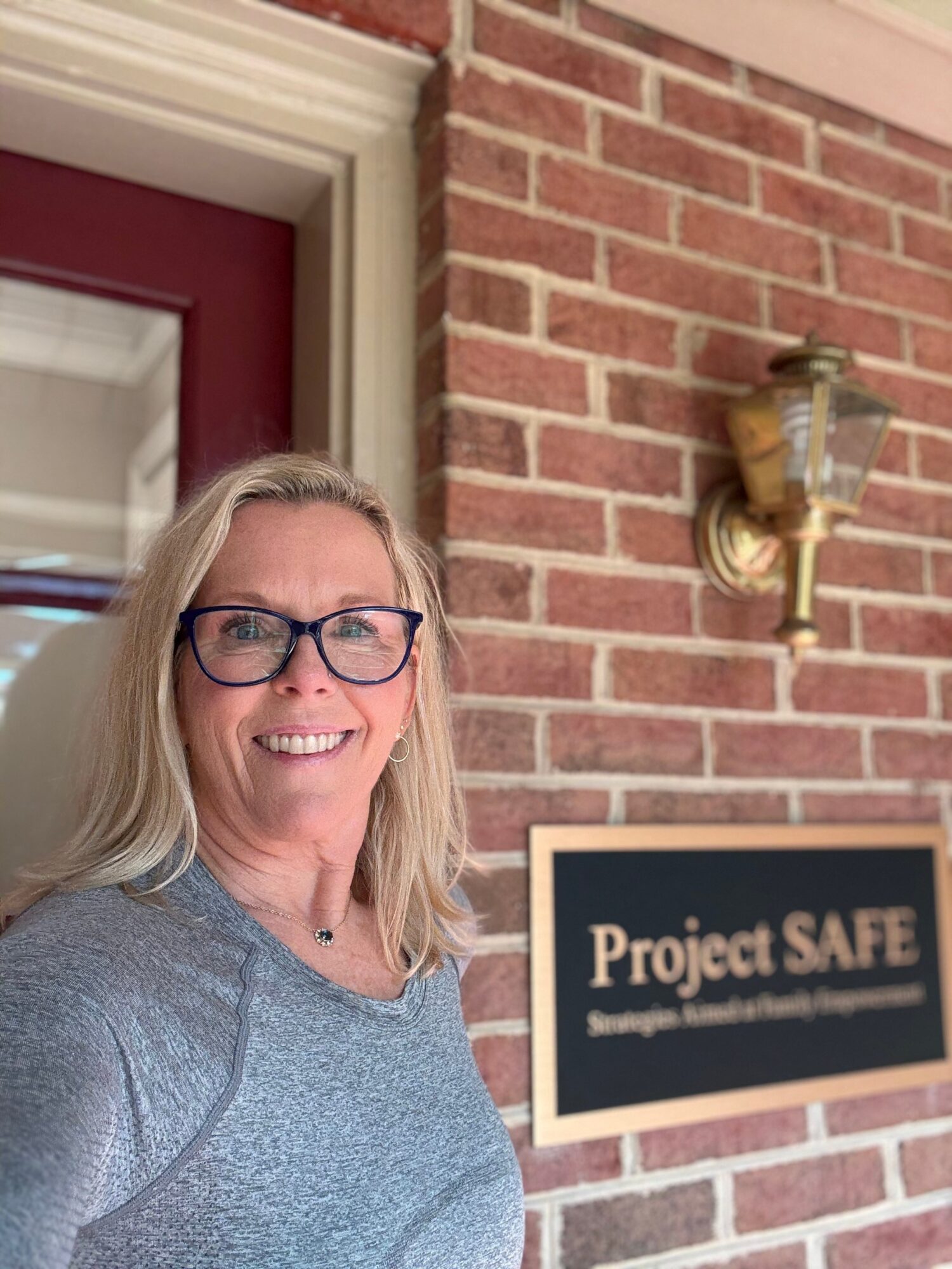 Woman with glasses smiling in front of a brick wall and a sign that reads 'Project SAFE'.