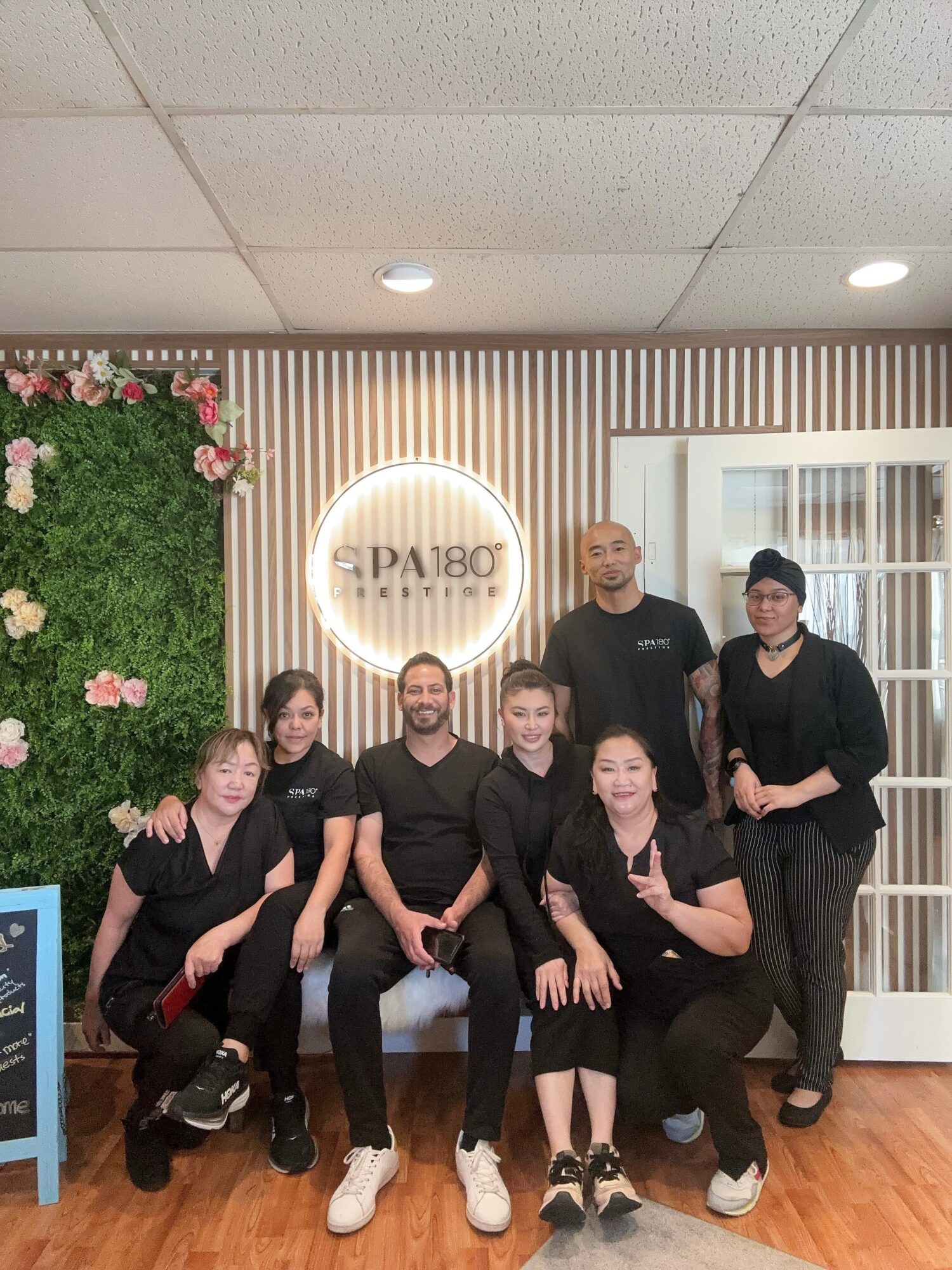 Group of seven people in black uniforms standing and sitting in a spa or salon, with a green plant and a circular sign in the background.