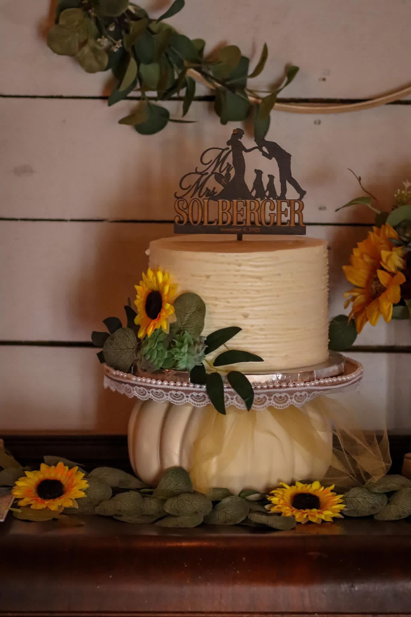 Cake with sunflower decorations, greenery, and a topper with a dog silhouette and text, on a wooden surface.