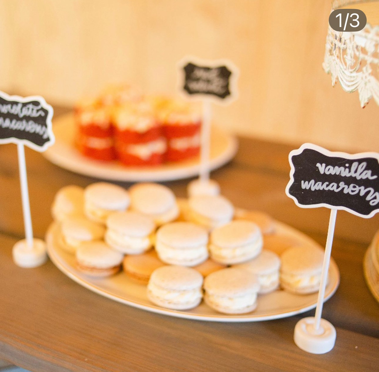 Plate of white macarons and red layered desserts on a wooden table, with chalkboard signs labeling the treats.