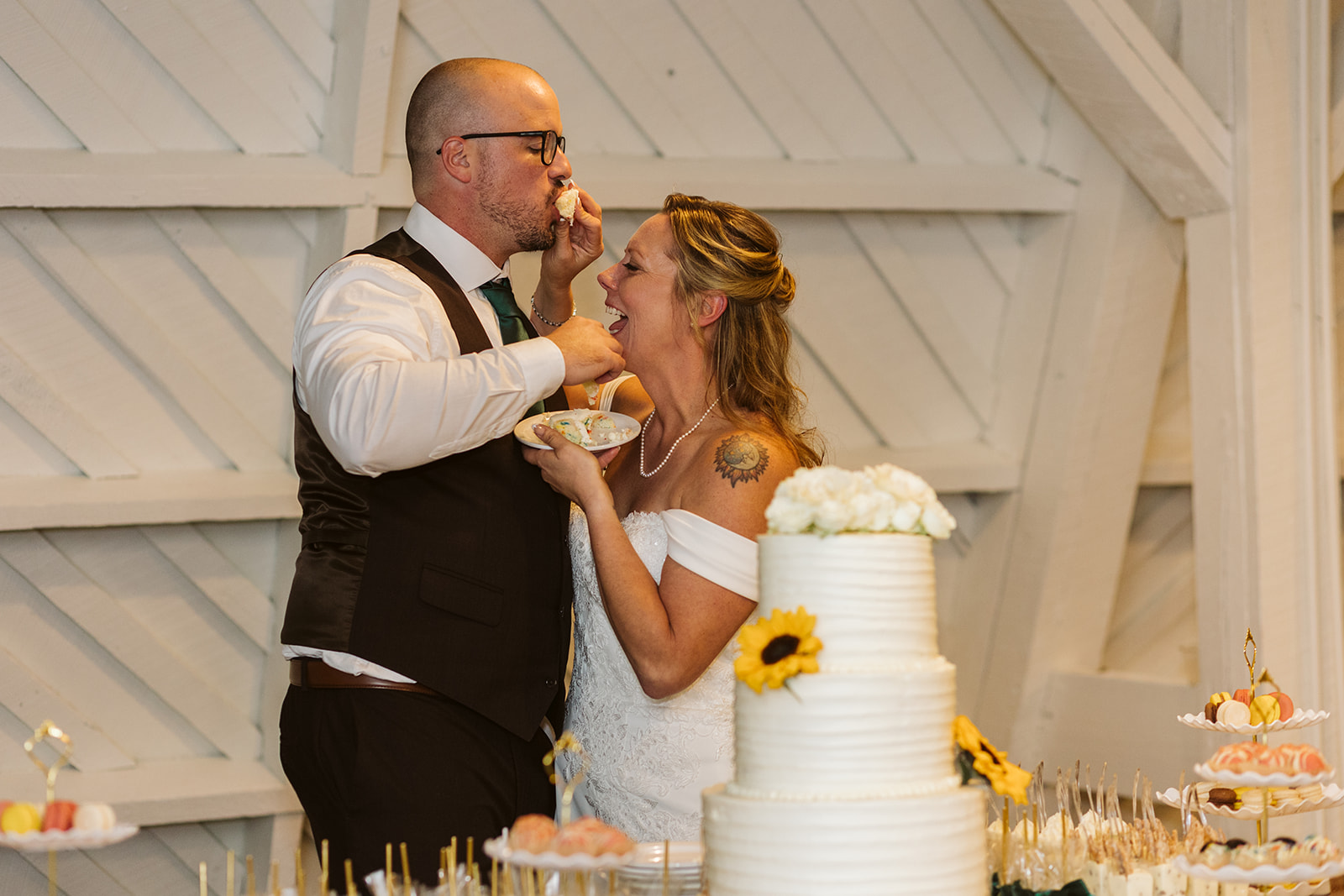 A couple in wedding attire sharing a moment indoors with a white wooden background, decorated with flowers and a cake.