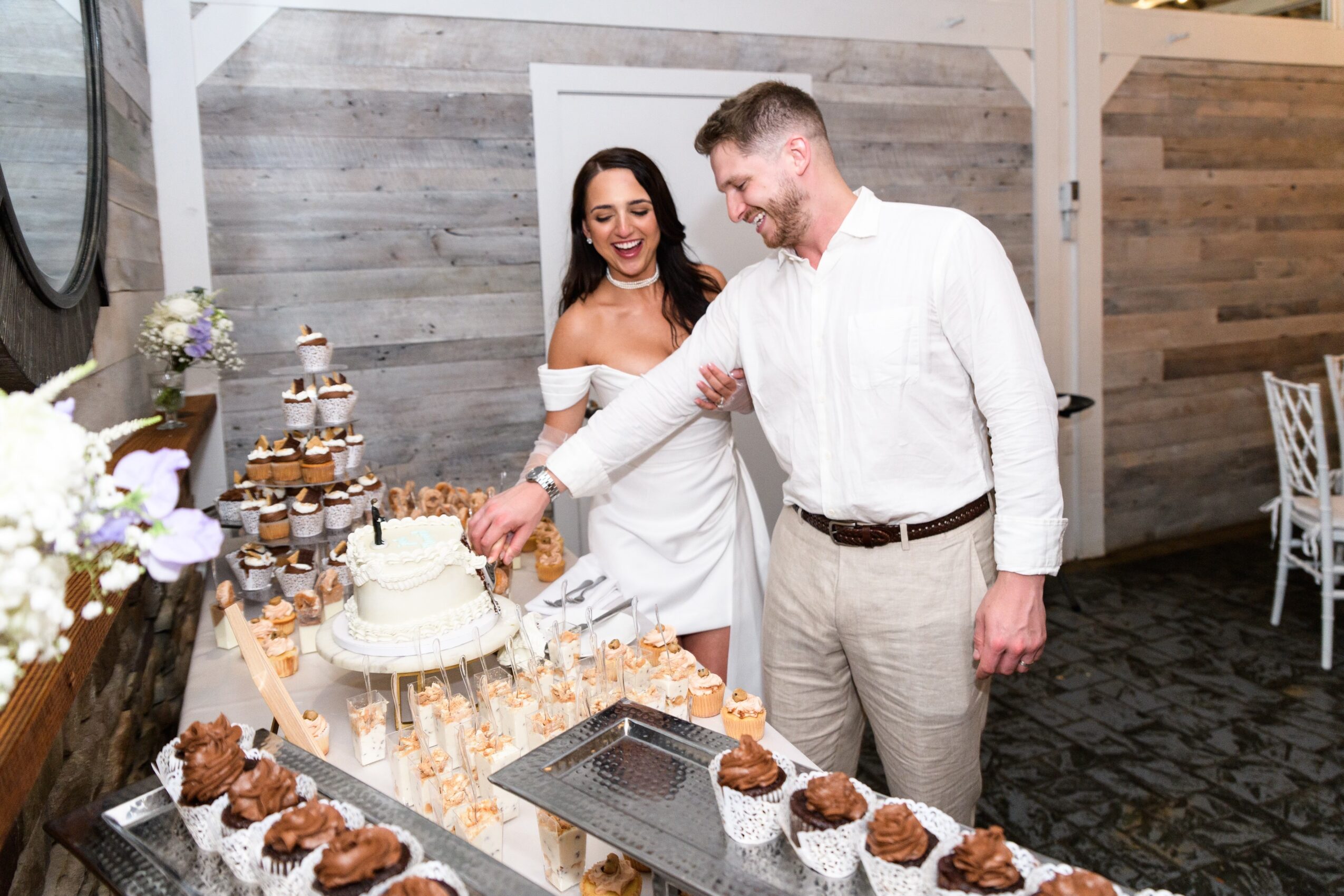 A man and woman cut a cake at a celebration, with cupcakes and desserts on the table, in a decorated room.