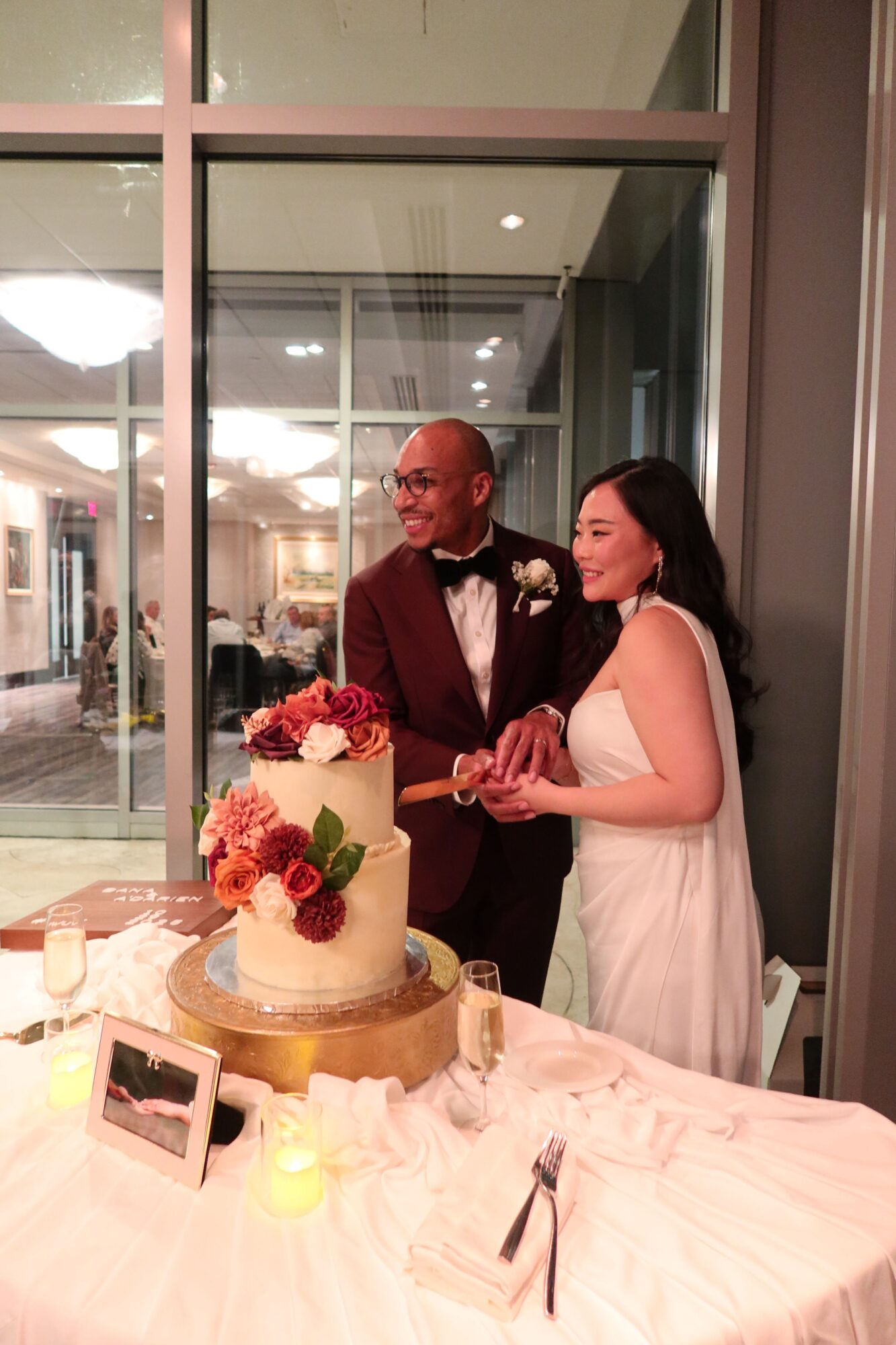 A man and woman are at a table with a cake and flowers, sharing a kiss in a modern indoor setting.