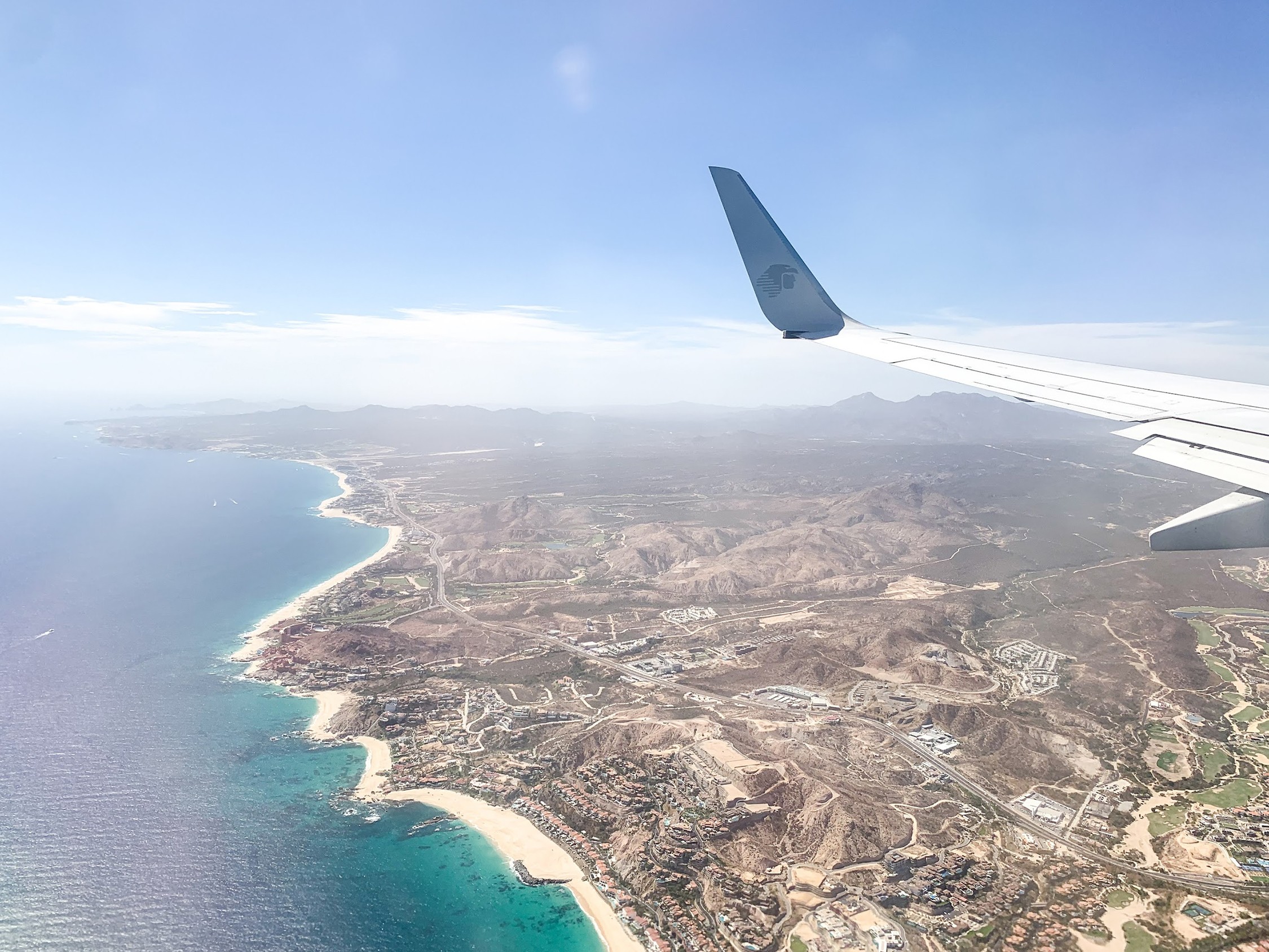 View from airplane window showing coastline, ocean, and land below with airplane wing visible.