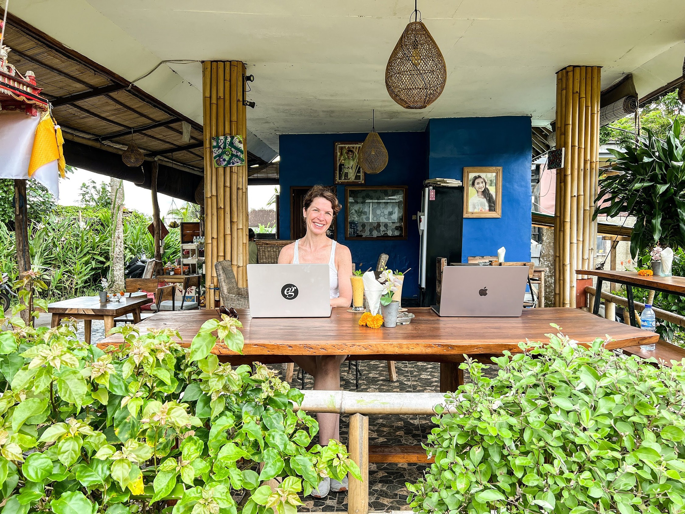 Woman sitting at a table with laptops, in an open-air restaurant with greenery, blue walls, and hanging light fixtures.