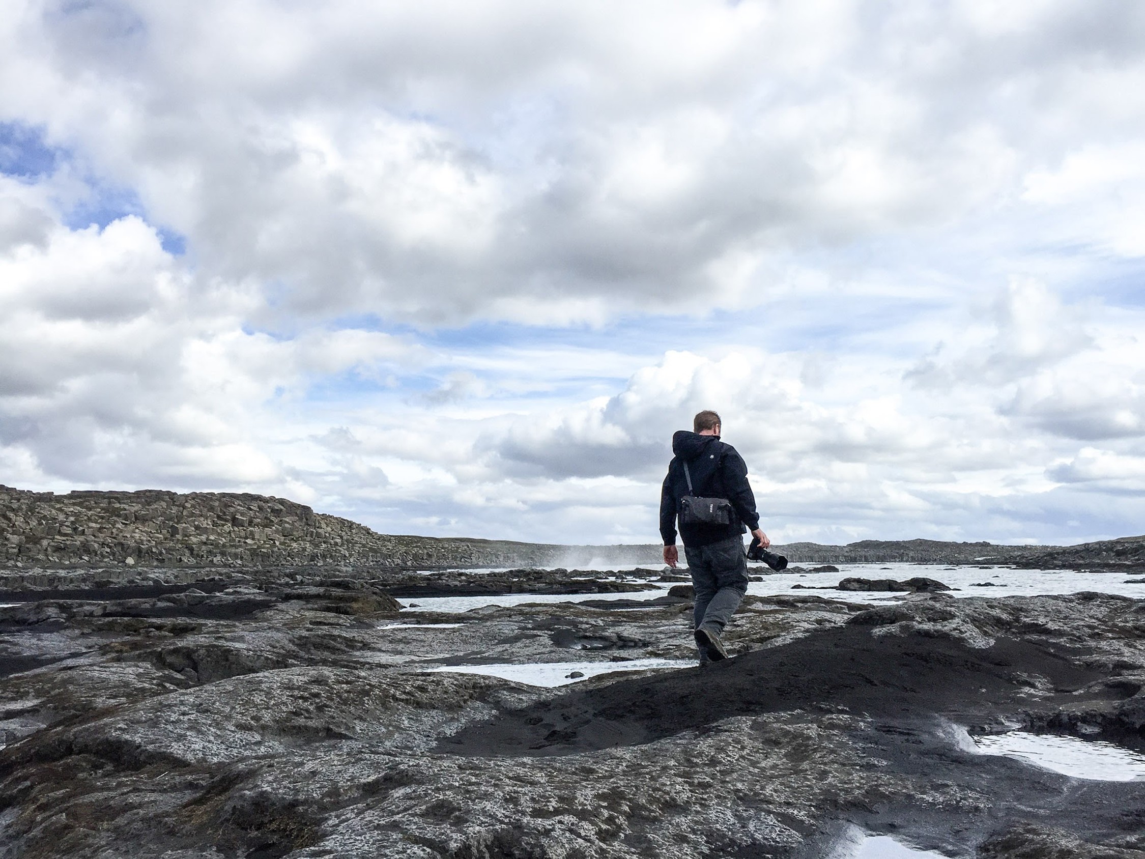 Person walking on rocky shoreline with cloudy sky and water in background.