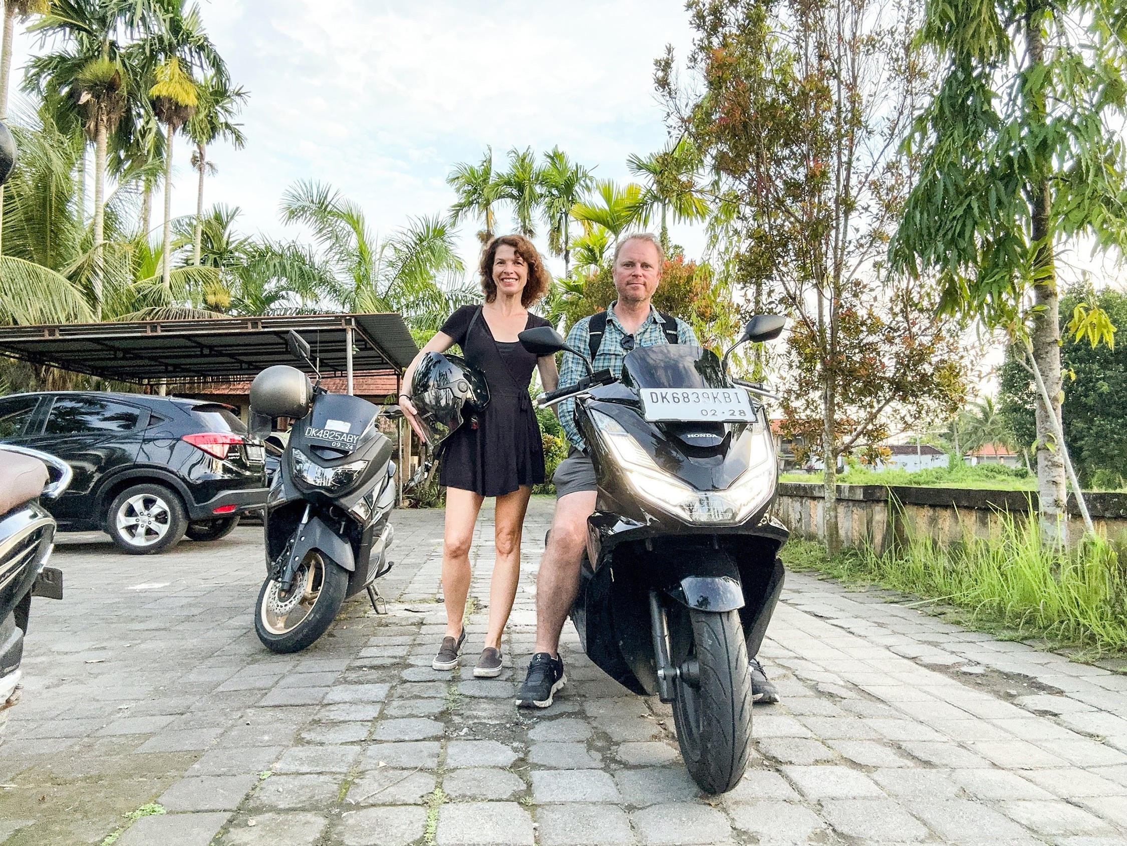 Two people standing next to scooters in a parking lot with trees and cars in the background.