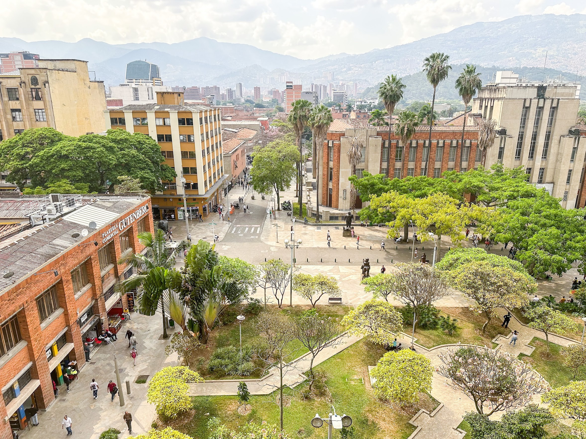 City park with trees, pathways, and buildings in the background, under a cloudy sky.