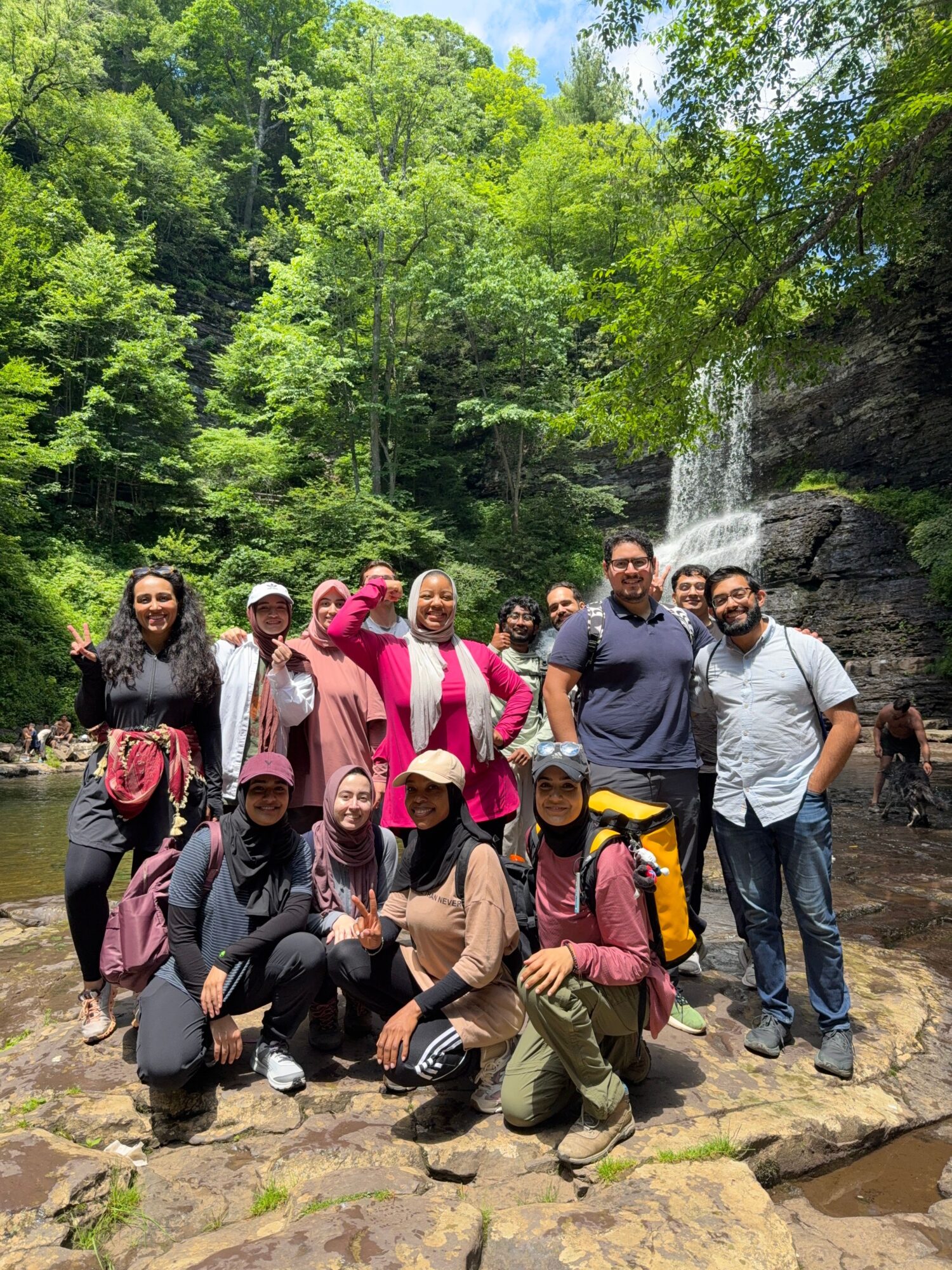 Group of ten people standing and kneeling near a waterfall in a forested area, smiling and posing for a photo.
