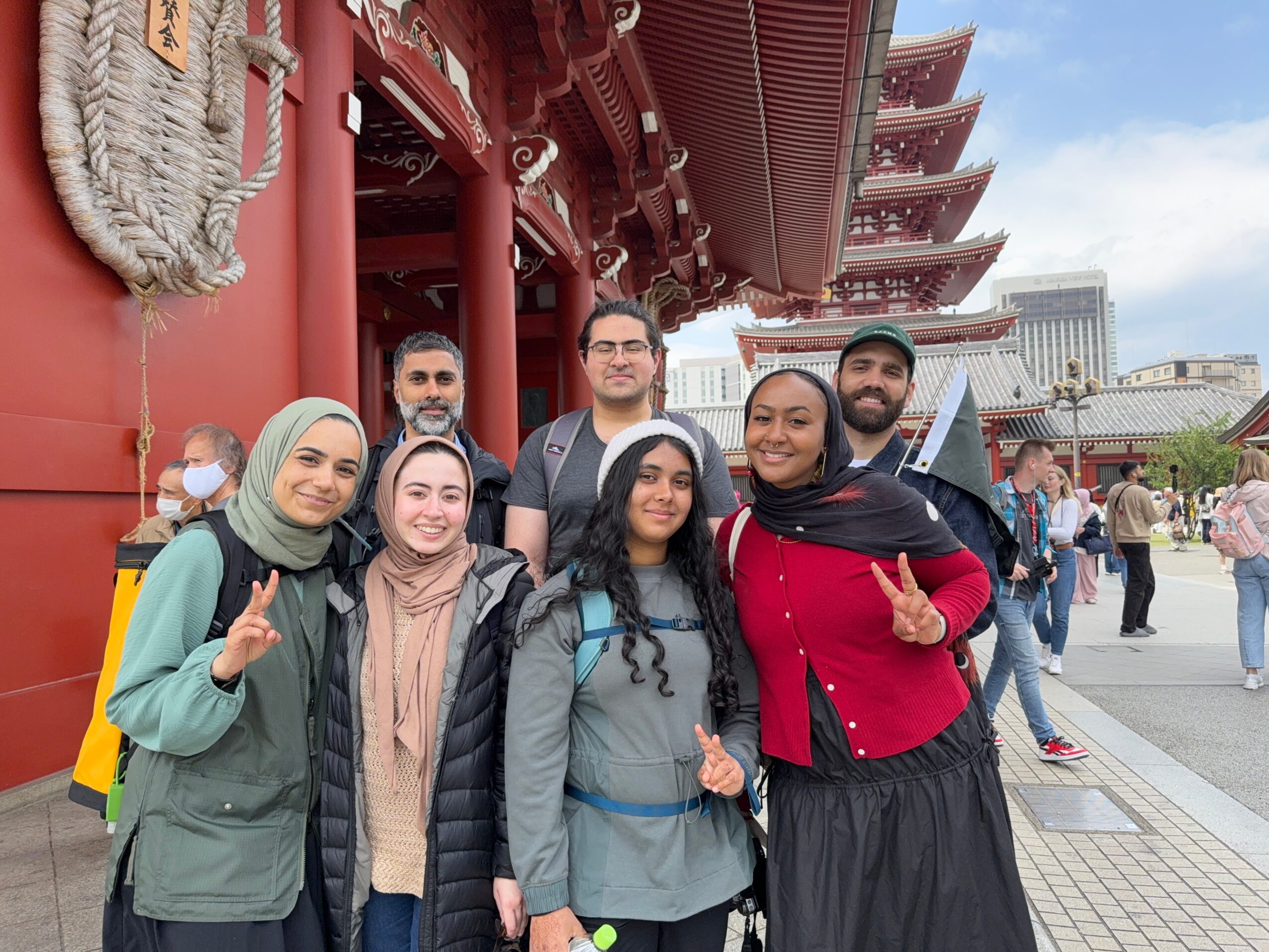 Group of seven diverse people standing in front of a traditional Asian temple with a pagoda-style roof, smiling and making peace signs.
