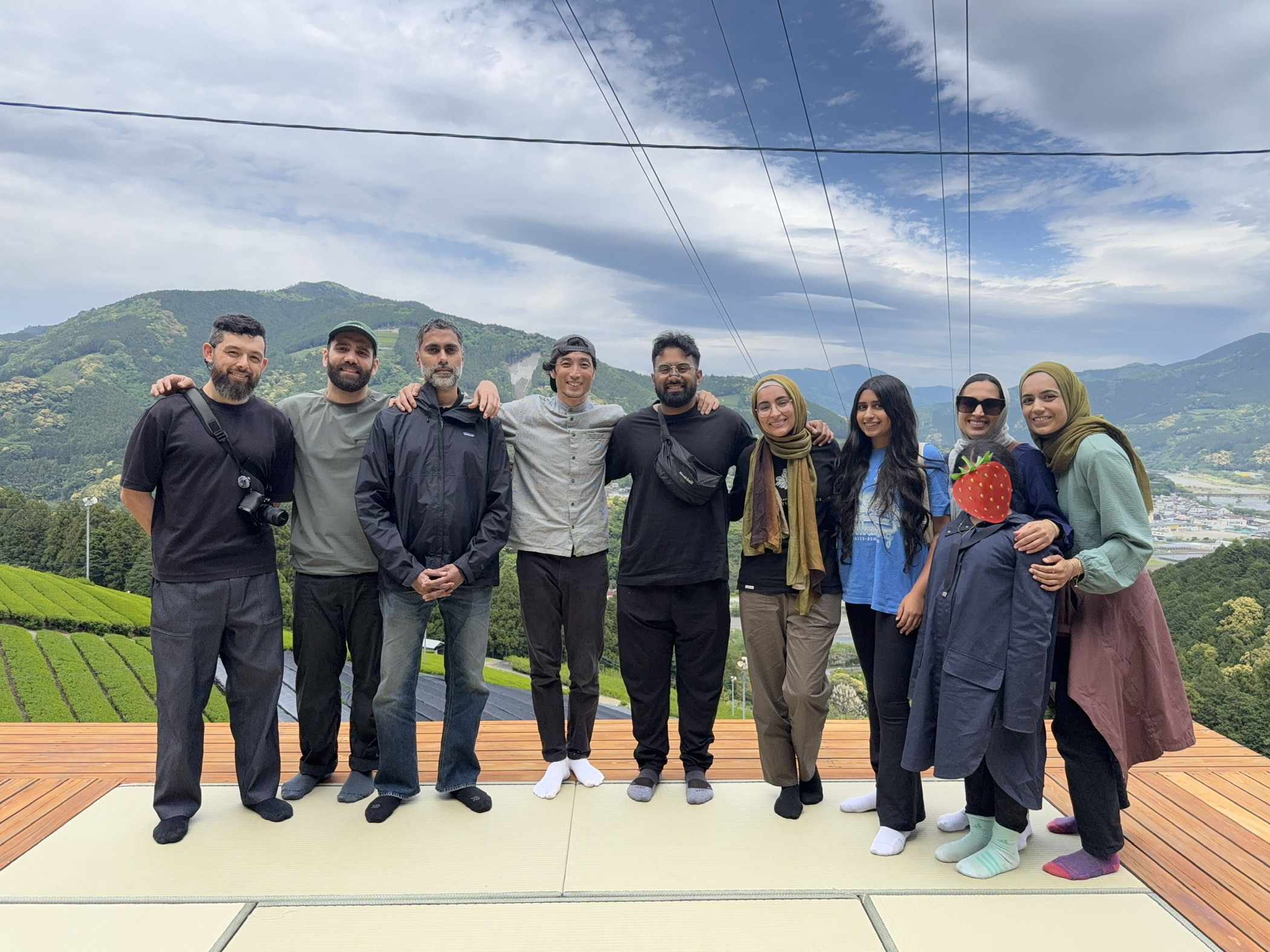 Group of nine people standing outdoors with mountains and sky in background, smiling and posing for photo.