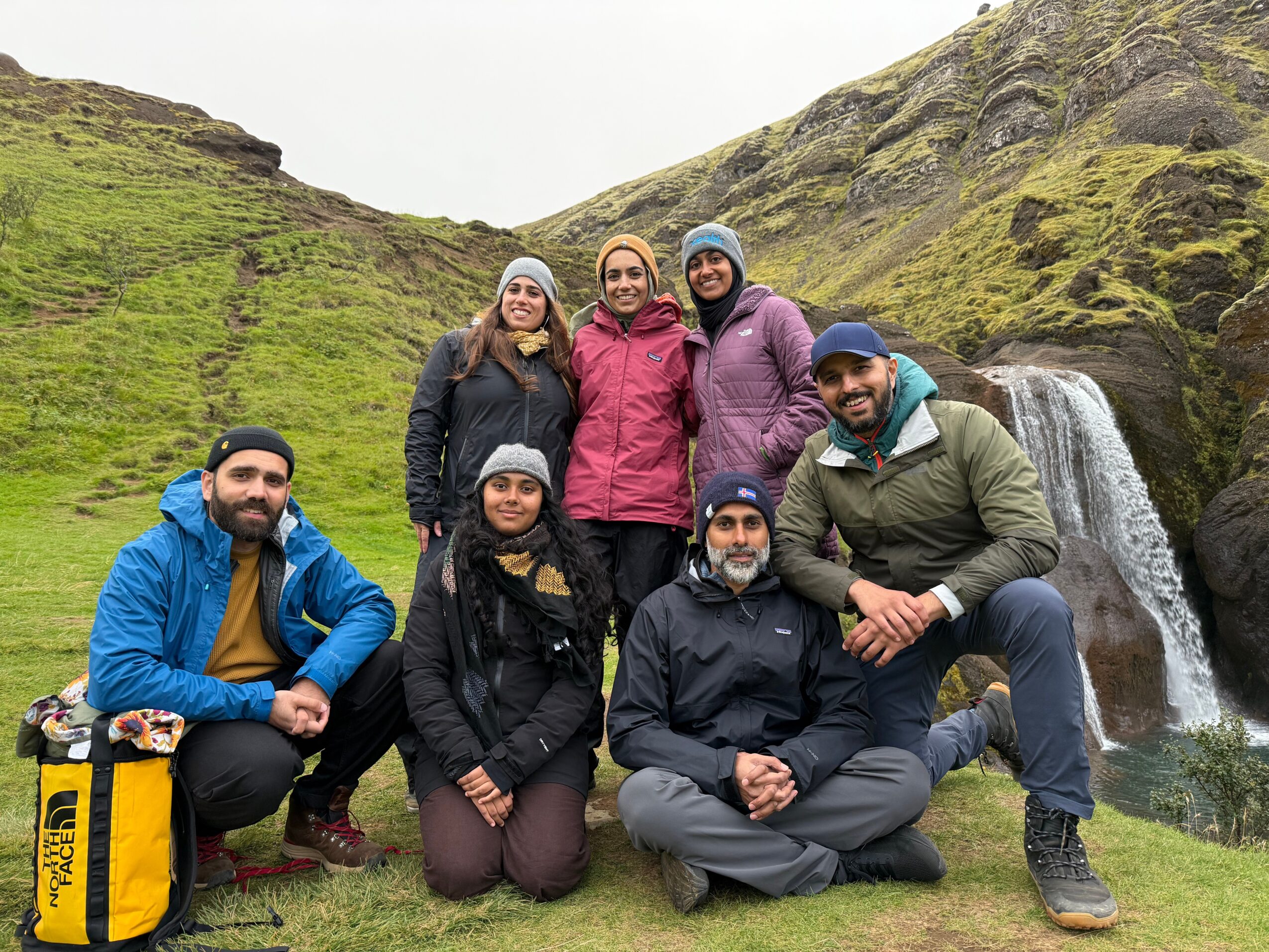 Group of seven people outdoors near a waterfall and green hills, posing for a photo, some wearing jackets and hats.