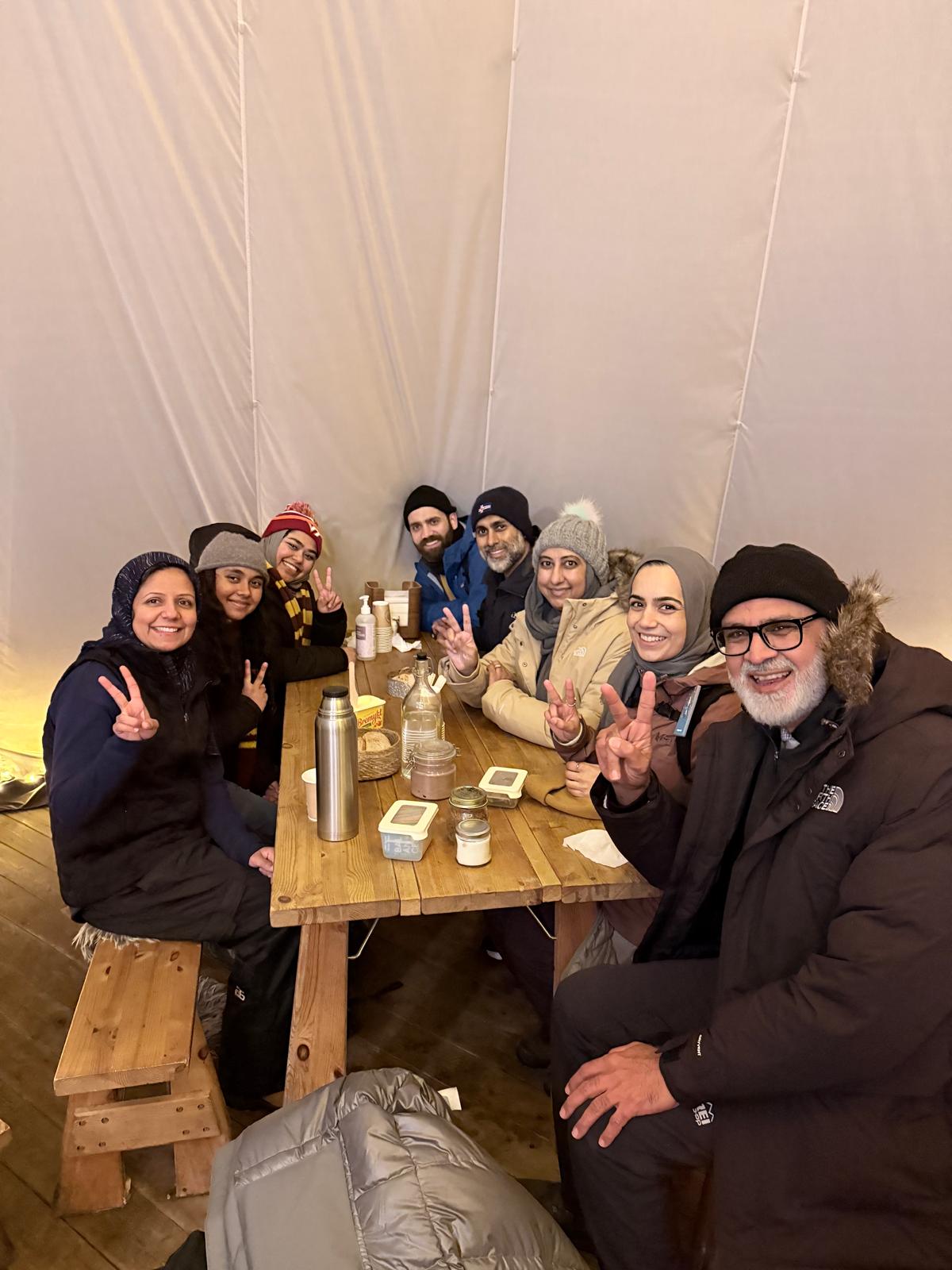 Group of eight people sitting at a wooden table inside a tent, smiling and making peace signs, with drinks and snacks.